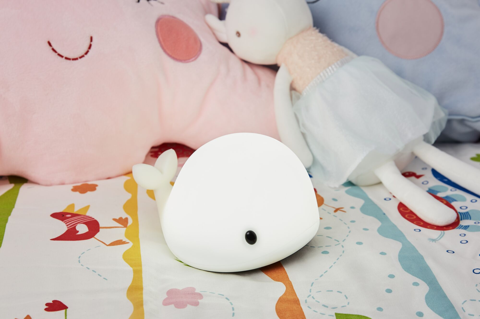 Białe nocne światło w kształcie wieloryba na kolorowym kocyku obok pluszaków.
