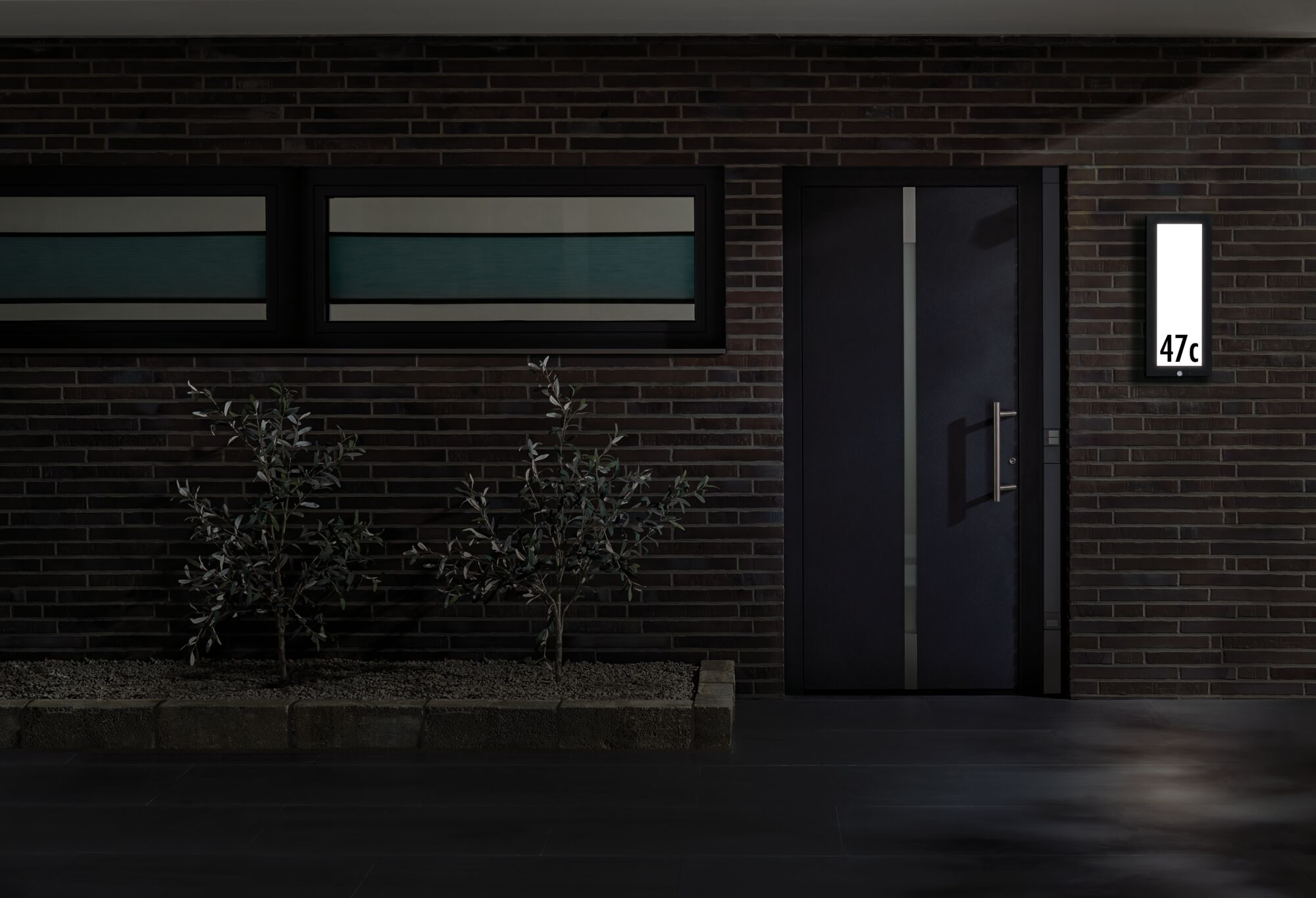 Ciemna ściana z cegły z drzwiami, dwoma oknami, lampą z numerem 47c i krzewami.