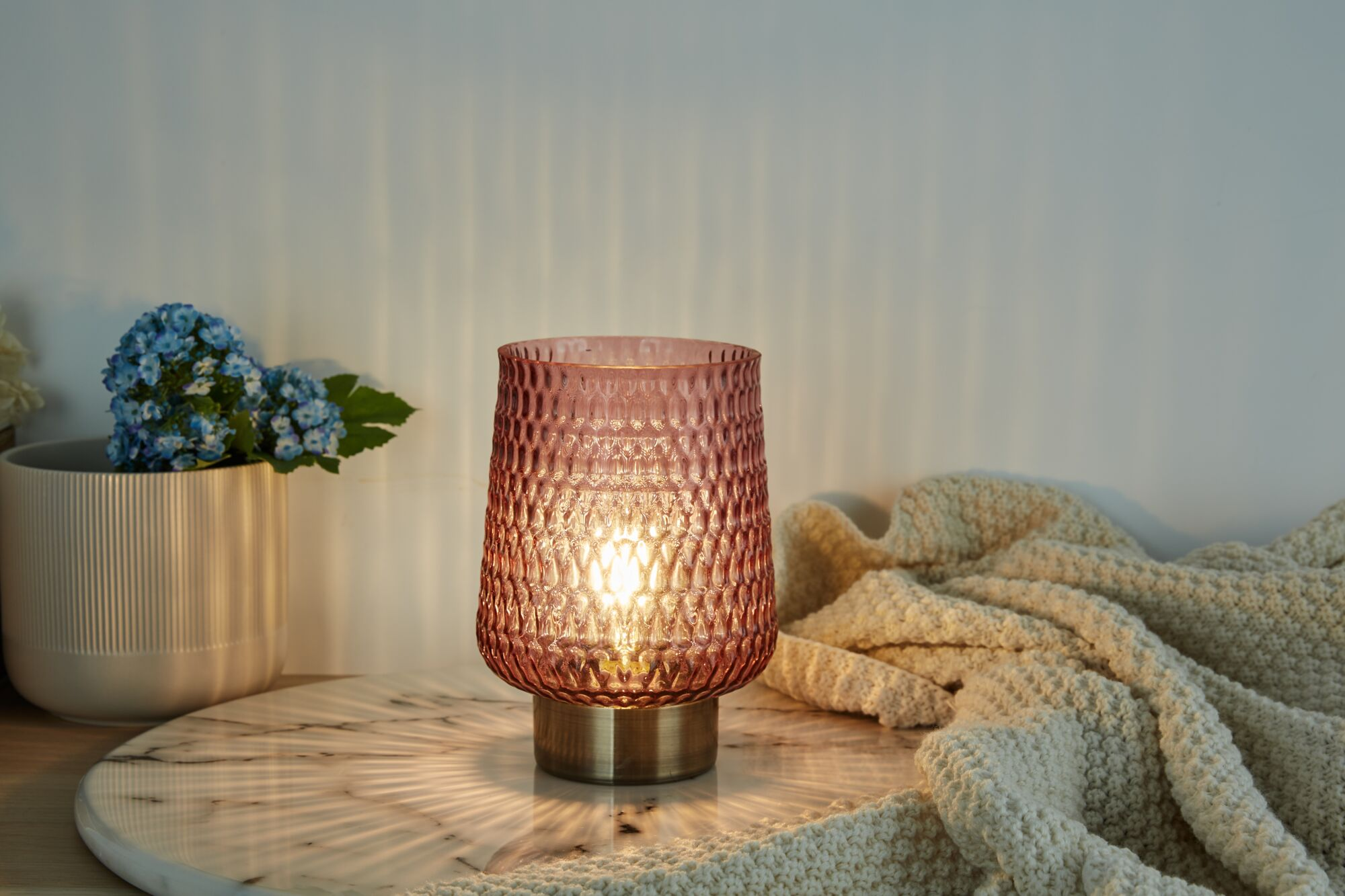 Różowa szklana lampa o teksturowanej powierzchni stoi na marmurowej tacy z niebieskimi kwiatami.