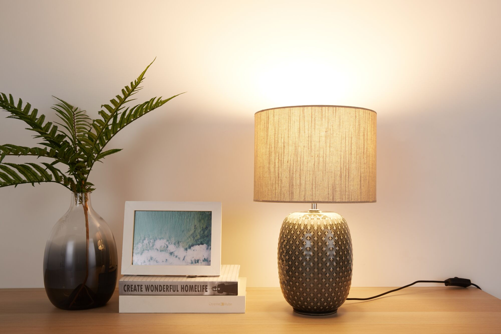 Lampa z wzorem, wazon z paprocią i ramka na zdjęcia na drewnianym stole.