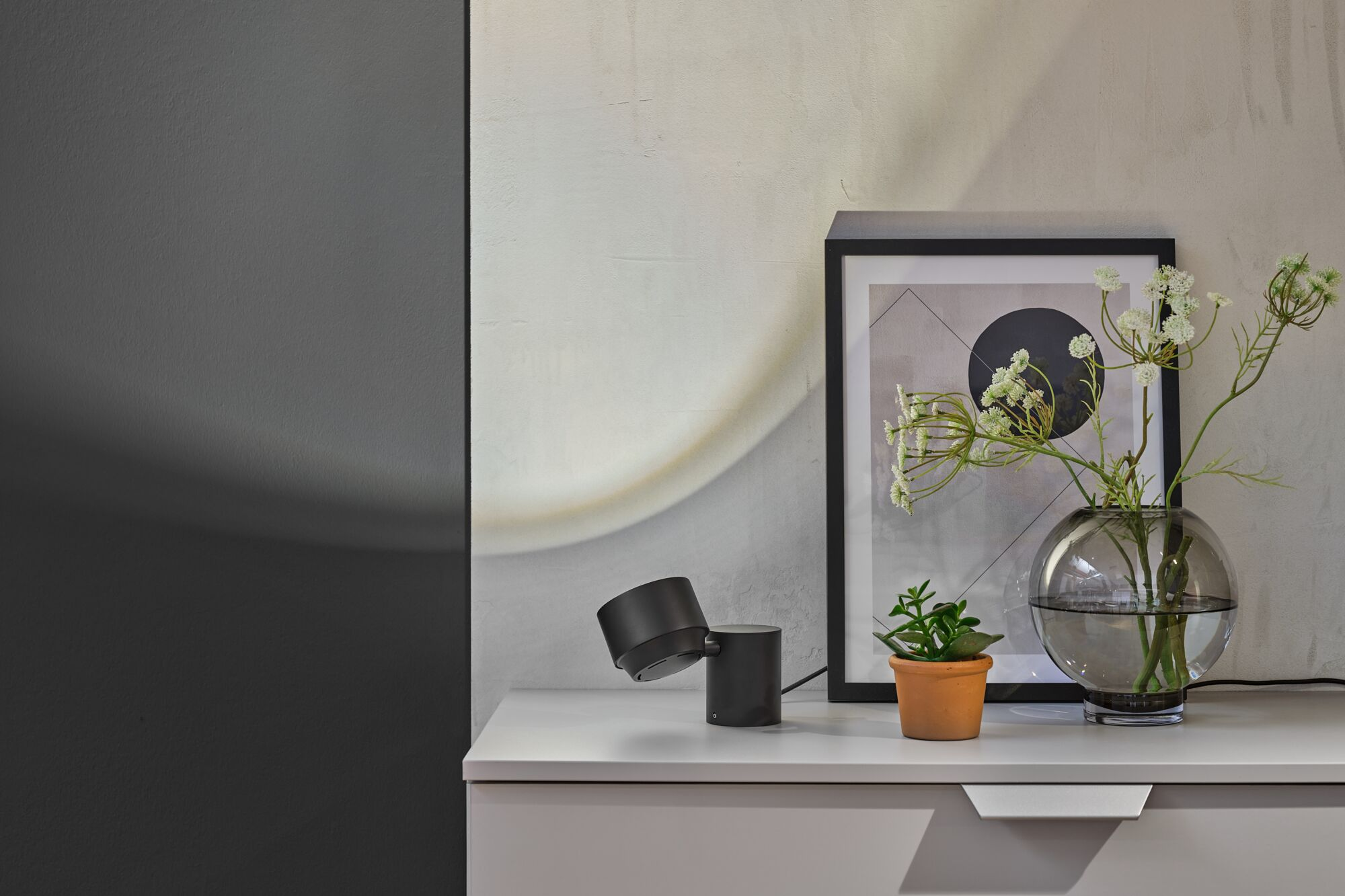Ein schwarzer Schreibtischlampenarm leuchtet auf einem weißen Regal neben einem gerahmten Bild und einer Vase.