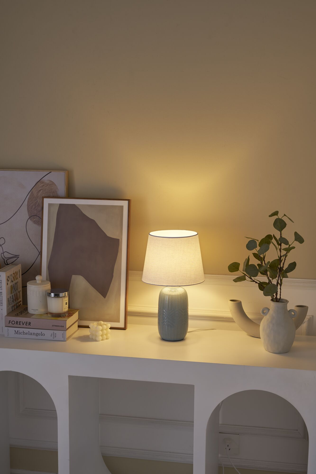 Lampa stołowa oświetla stolik boczny z książkami, świecami i sztuką.