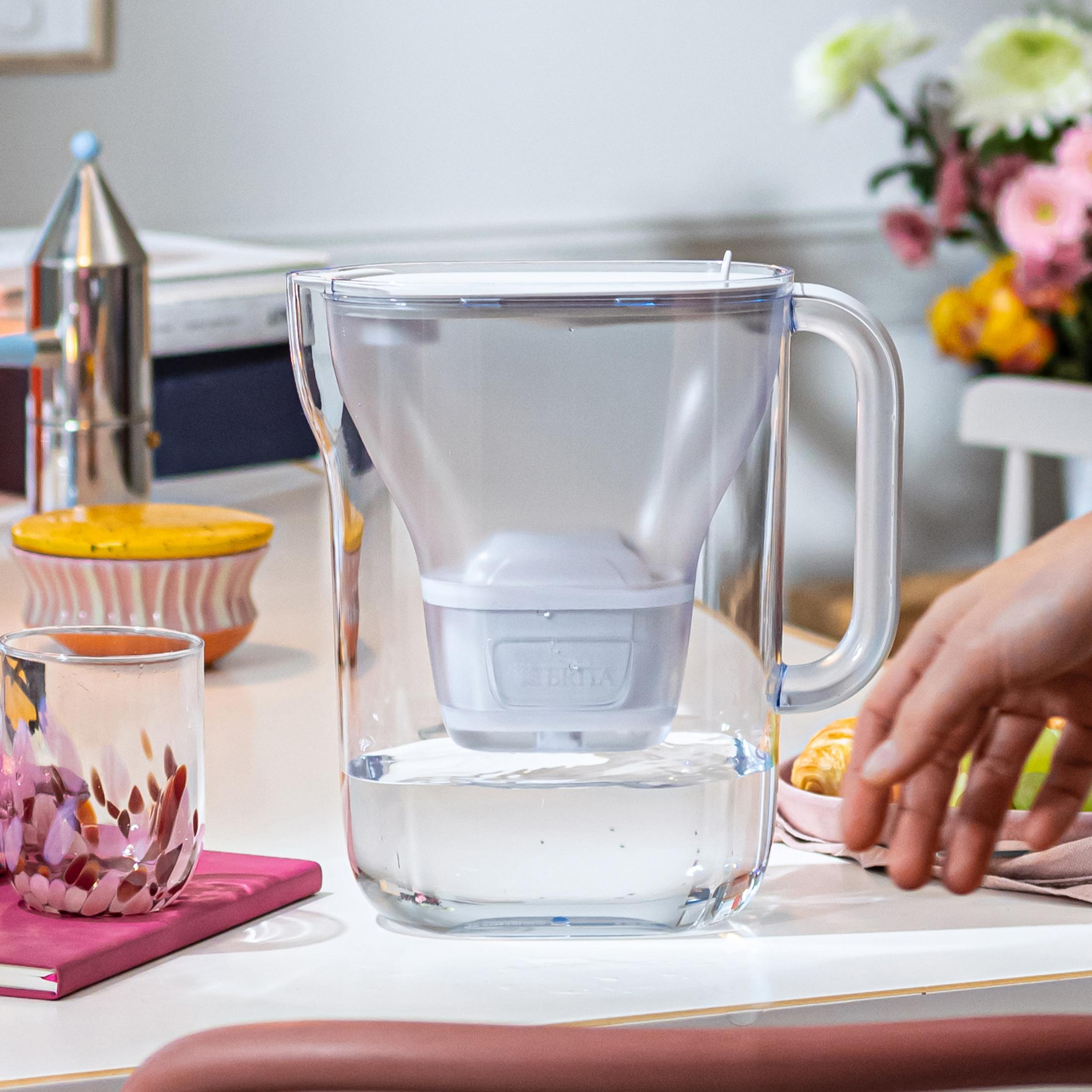 Una caraffa filtrante Brita trasparente con filtro su un tavolo. Vicino ci sono bicchieri e fiori.