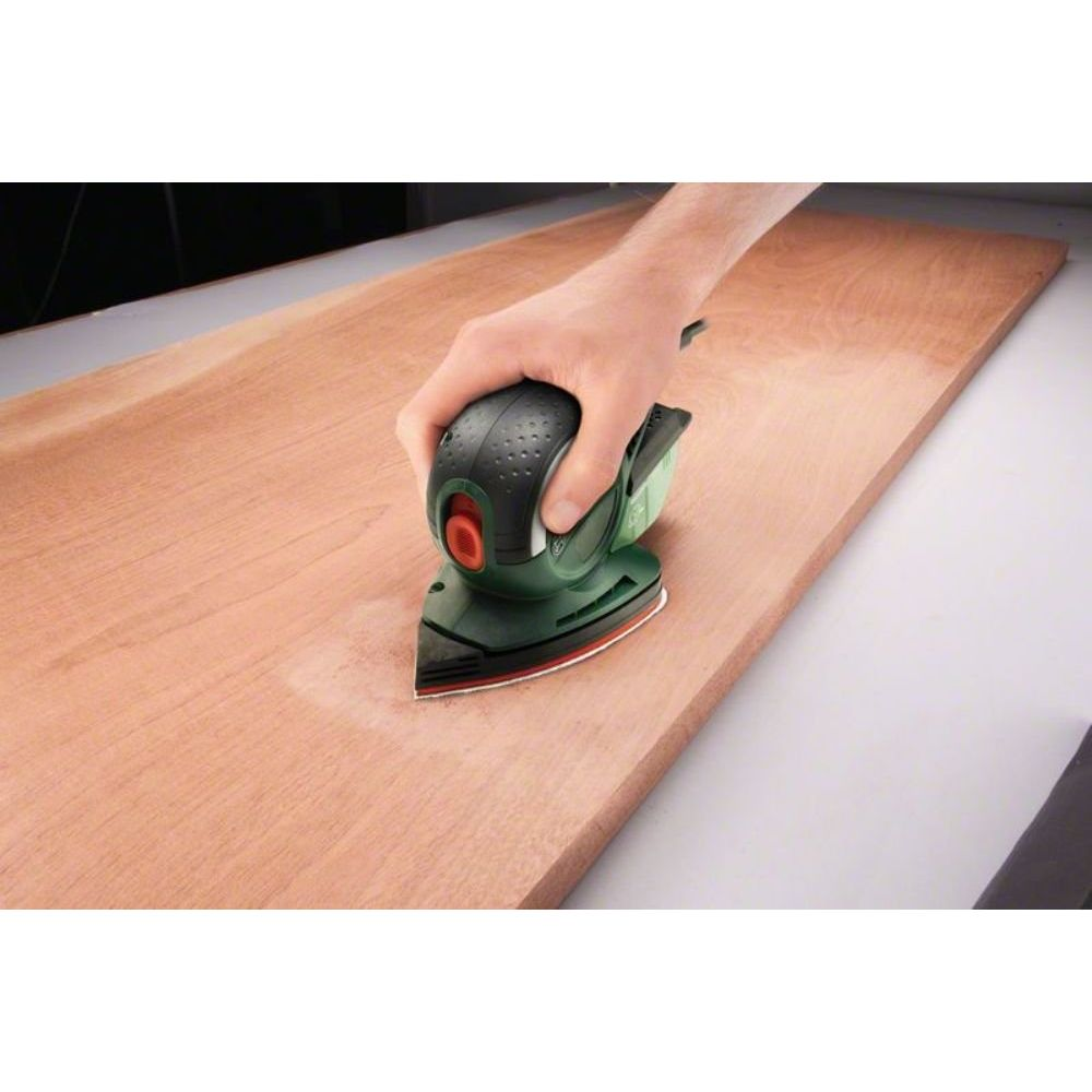 Eine Hand benutzt einen grünen Schwingschleifer auf einer Holzoberfläche. Staub ist sichtbar.
