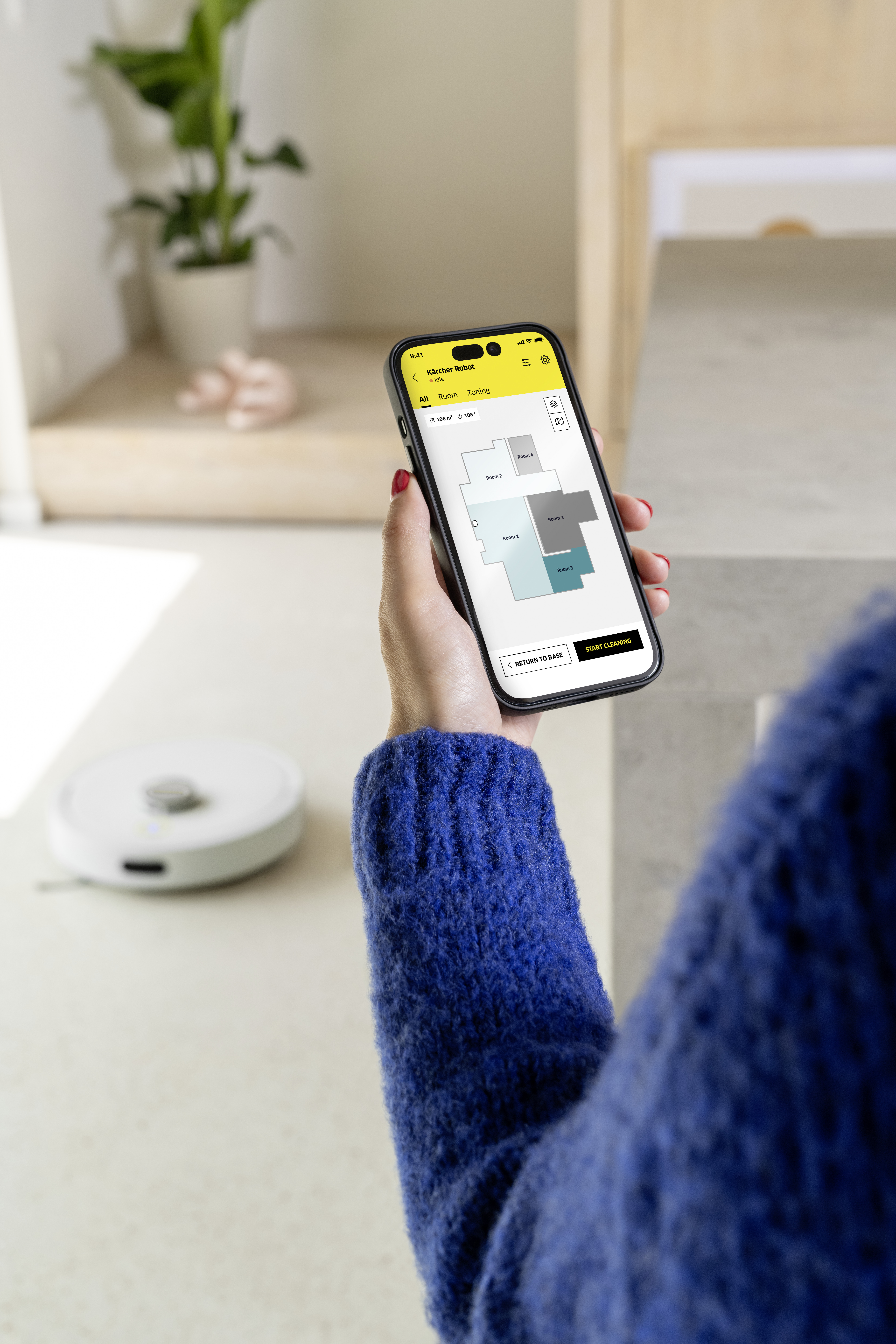 Osoba obsługuje aplikację Kärcher Robot na smartfonie. W tle widoczny jest robot sprzątający.