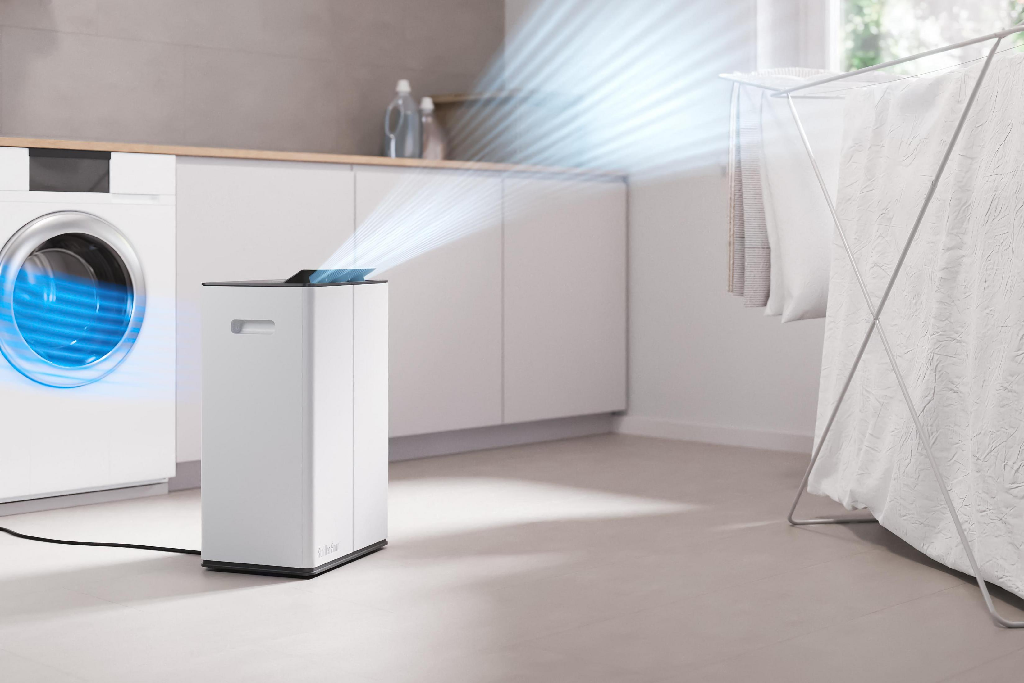 Een witte luchtreiniger zendt blauwe luchtstromen uit, naast een wasmachine.