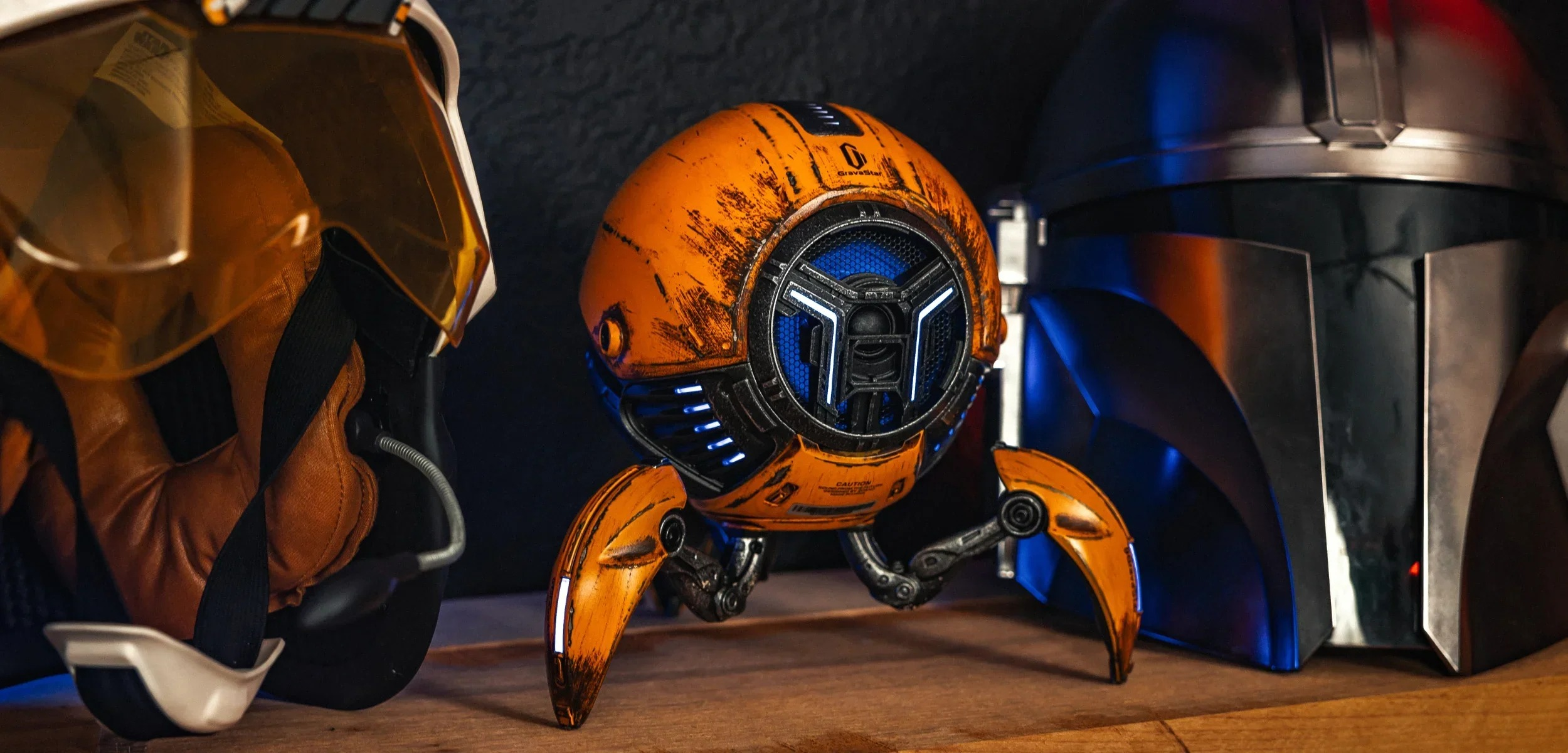 Pomarańczowy robot z niebieskimi światłami stoi między dwoma hełmami.