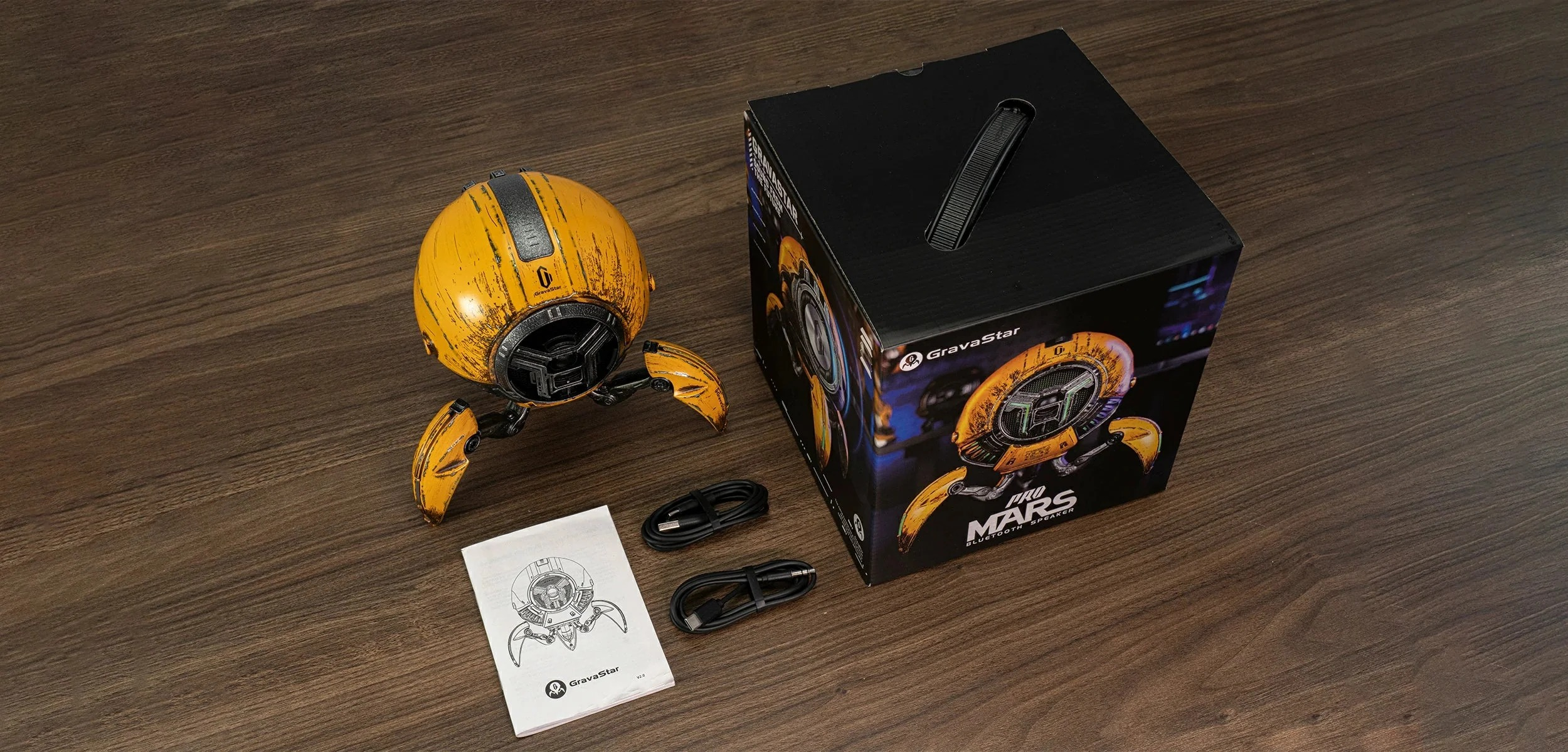 Żółty głośnik-robot z nogami, opakowaniem i kablami na drewnianej podłodze.