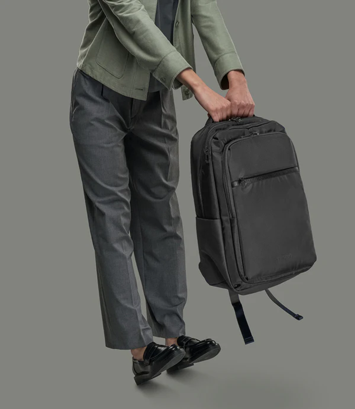 Una persona con pantaloni grigi, giacca verde e scarpe nere solleva uno zaino nero.