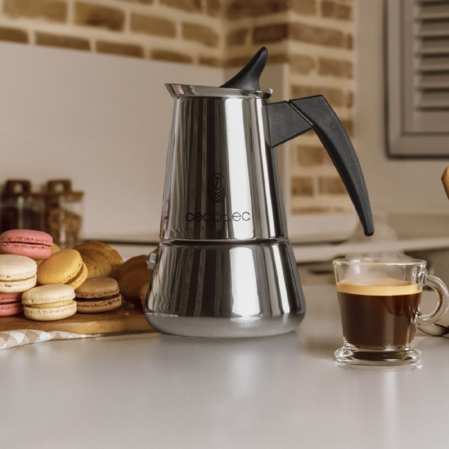 Una caffettiera espresso argentata accanto a un bicchiere di caffè e macarons su un piano di lavoro.