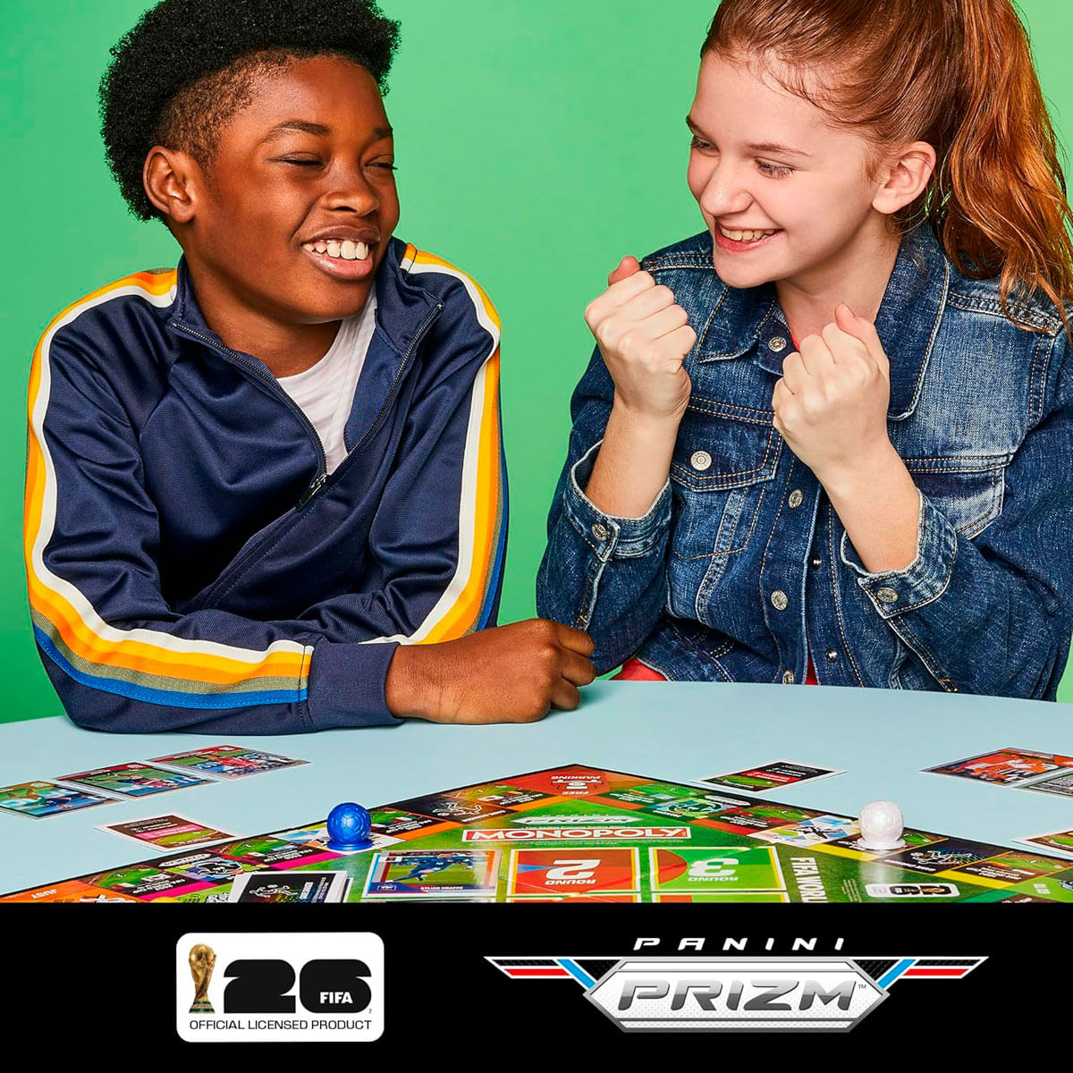 Dos niños riendo juegan al Monopoly con cartas de fútbol sobre fondo verde.