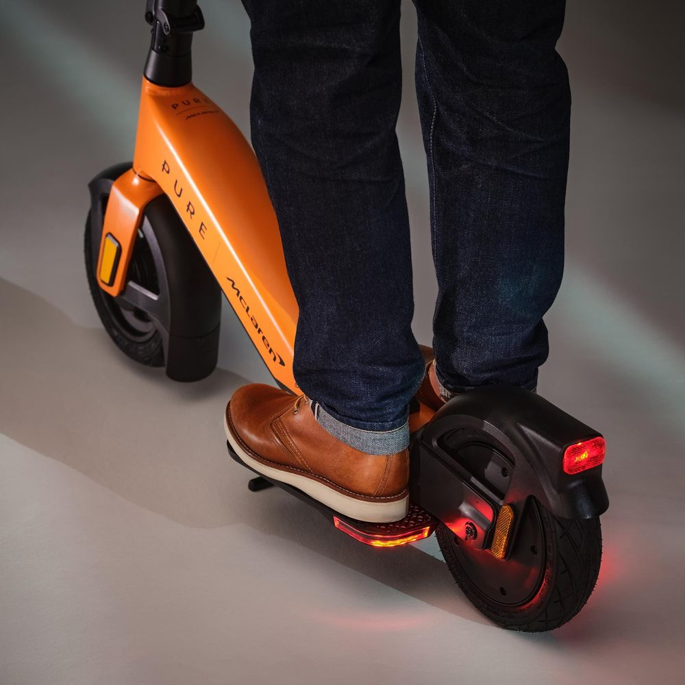Person auf orangefarbener McLaren E-Scooter, Jeans, braune Schuhe. Rotes Rücklicht.