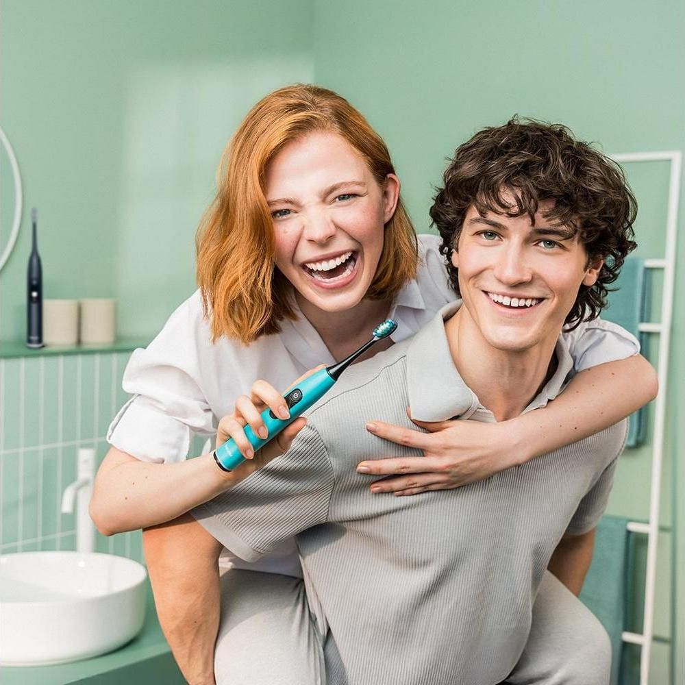 Een man en vrouw lachen in een badkamer, zij houdt een tandenborstel vast.
