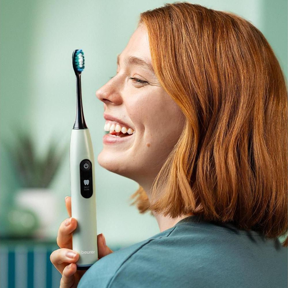 Smiling woman with red hair holds a white electric toothbrush with a blue and green brush head.