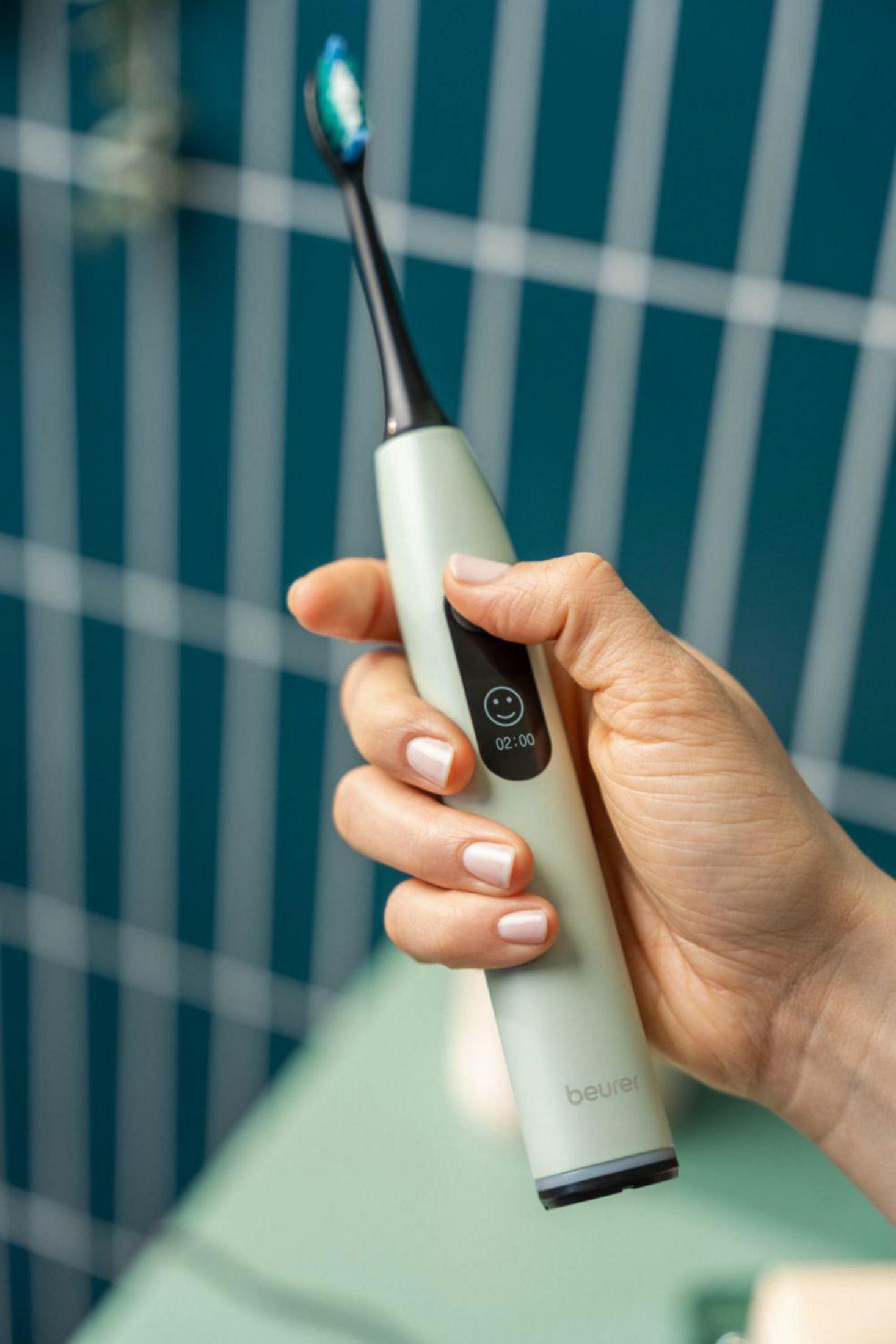 A hand holds a light green electric toothbrush with blue and white bristles.