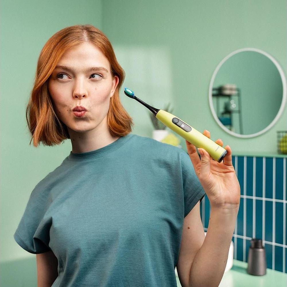 A woman with red hair holds a yellow electric toothbrush with blue bristles.