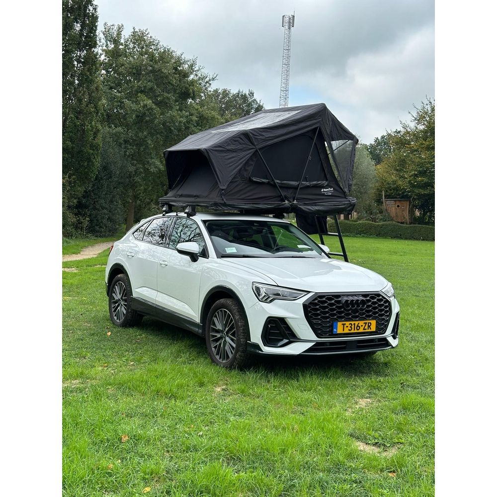 Un SUV Audi bianco con tenda sul tetto su un prato, con alberi e torre cellulare dietro.