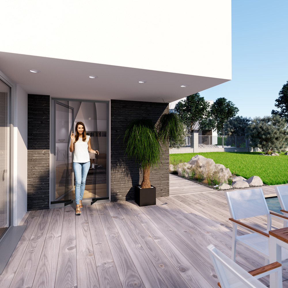 Eine Frau in Jeans und weißem Oberteil steht auf einer Holzterrasse vor einem modernen Haus.