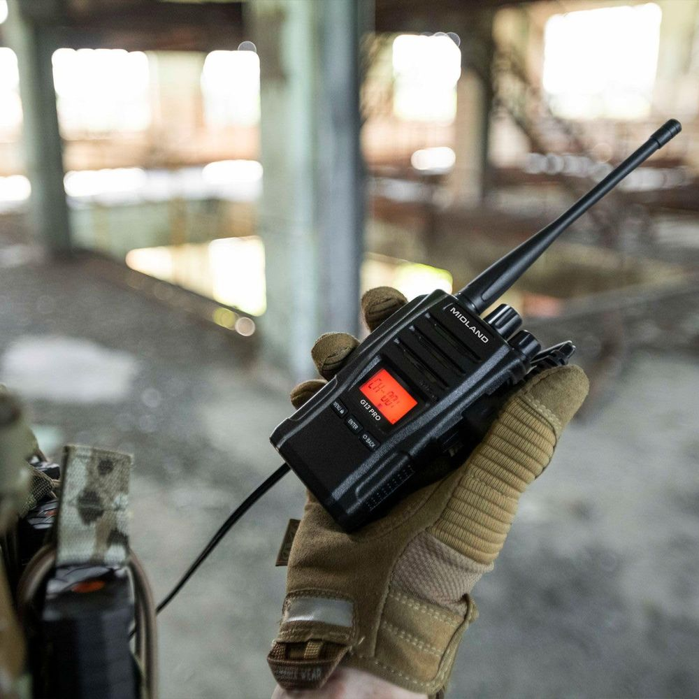 Una mano guantata tiene un walkie-talkie nero con display rosso in un edificio in rovina.