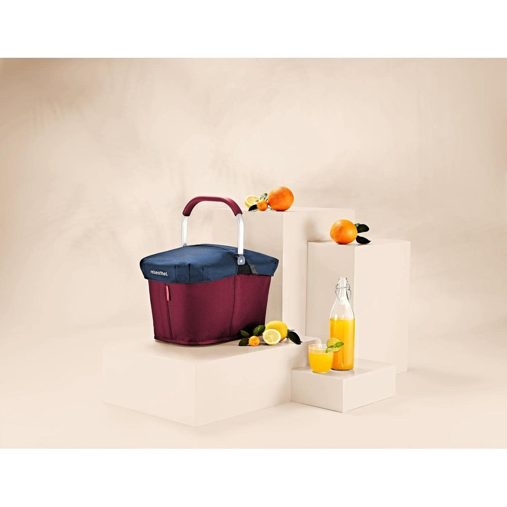 Eine bordeauxrote Einkaufstasche mit blauem Deckel steht neben Orangen und Saft.