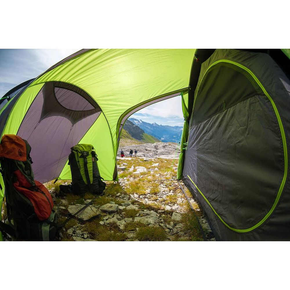 Interno di una tenda con zaini, affacciata su un paesaggio montano con escursionisti.