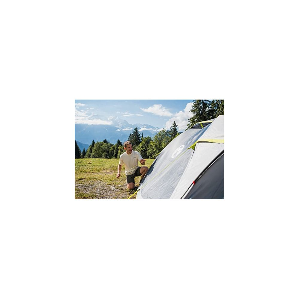 Un uomo si inginocchia accanto a una tenda grigia in un prato con montagne sullo sfondo.