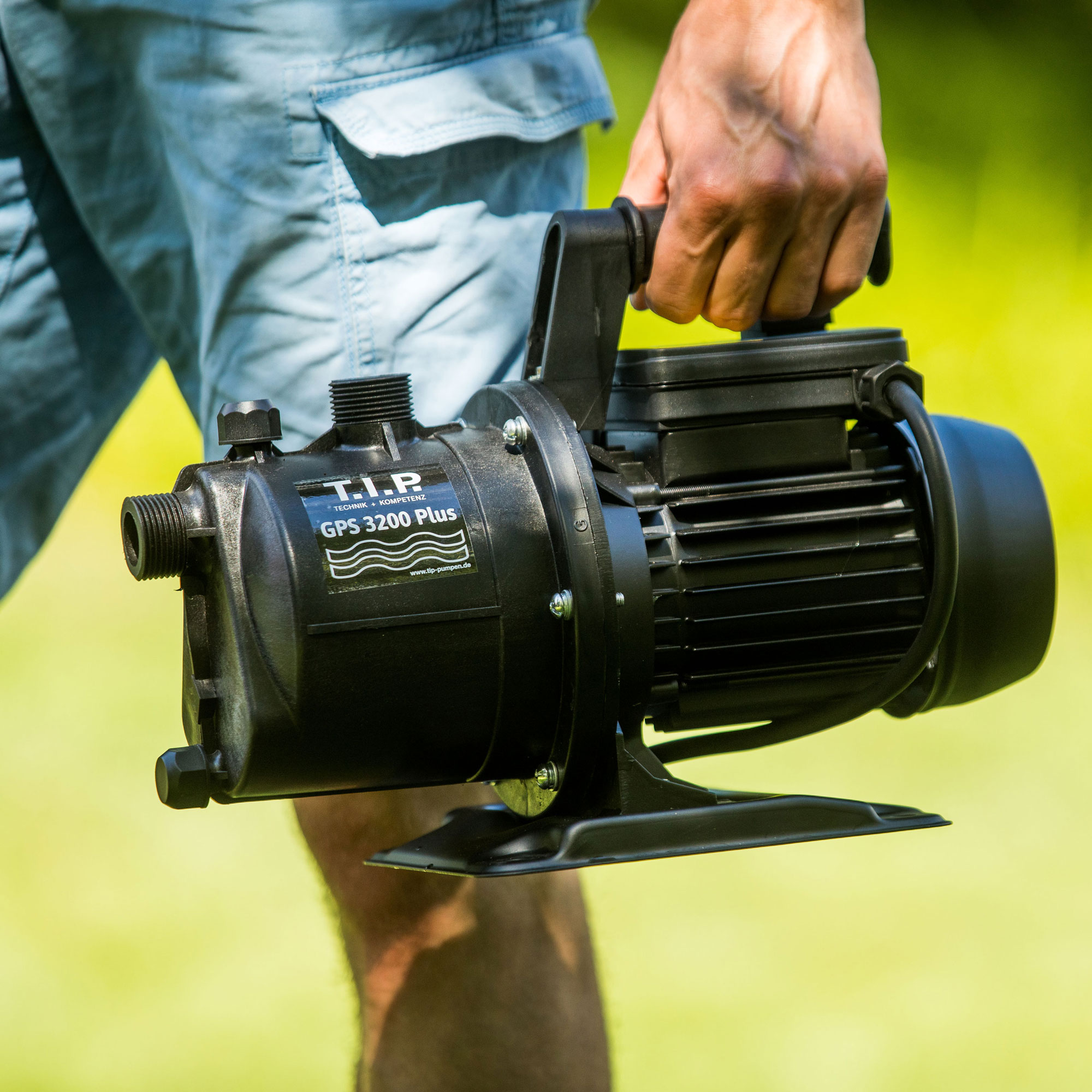 Eine Person trägt eine schwarze Wasserpumpe mit Griff vor unscharfem grünem Hintergrund.