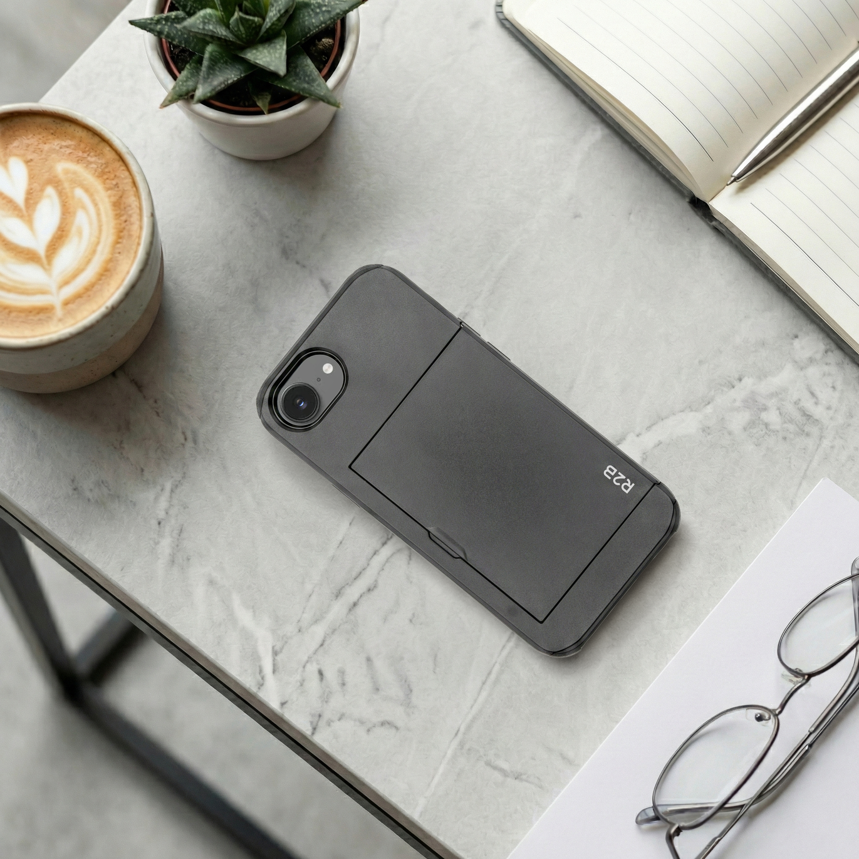 Donkere telefoon op marmeren tafel met koffie, plant, notitieboek en bril.