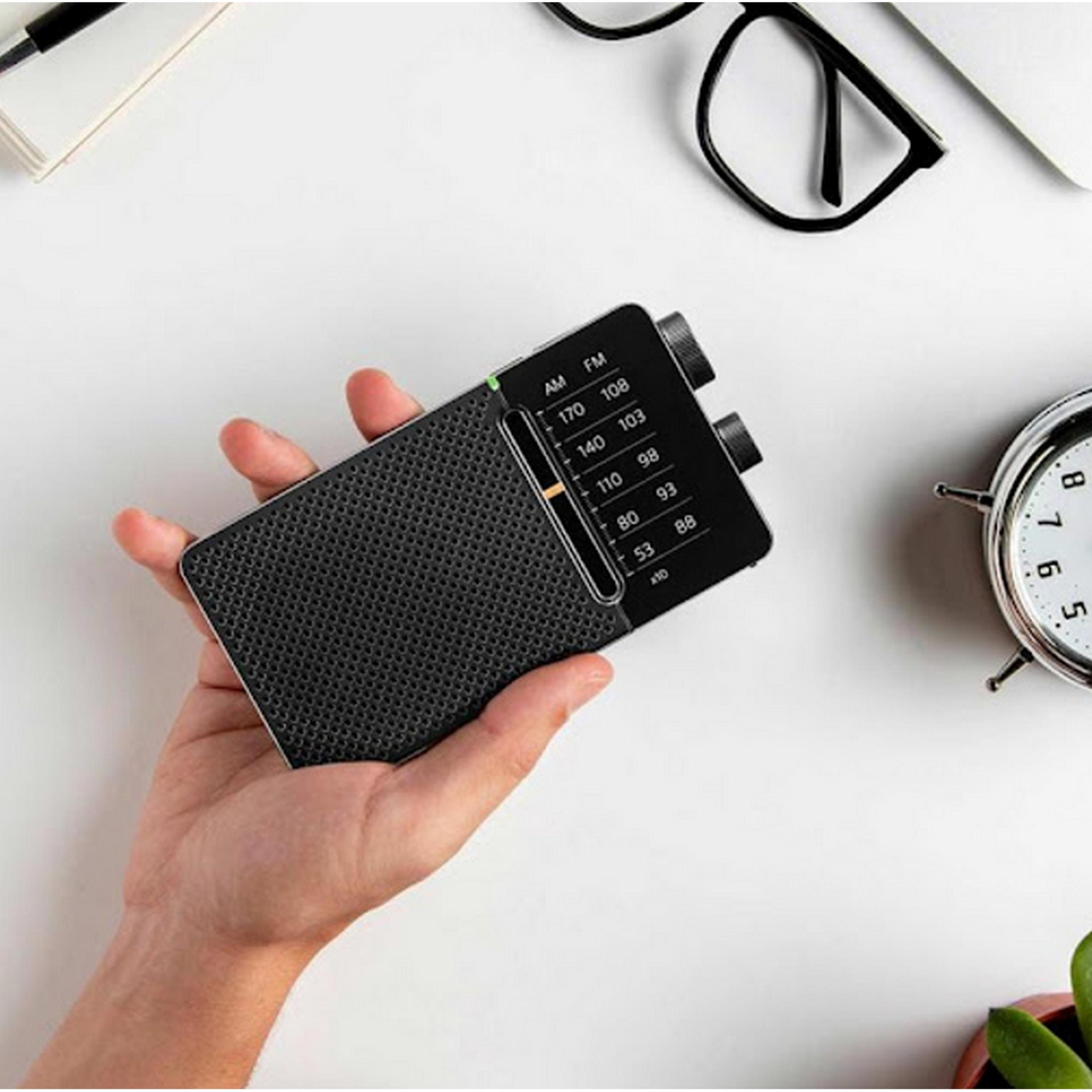 Hand holds a black radio with AM/FM dial. Clock and glasses nearby.