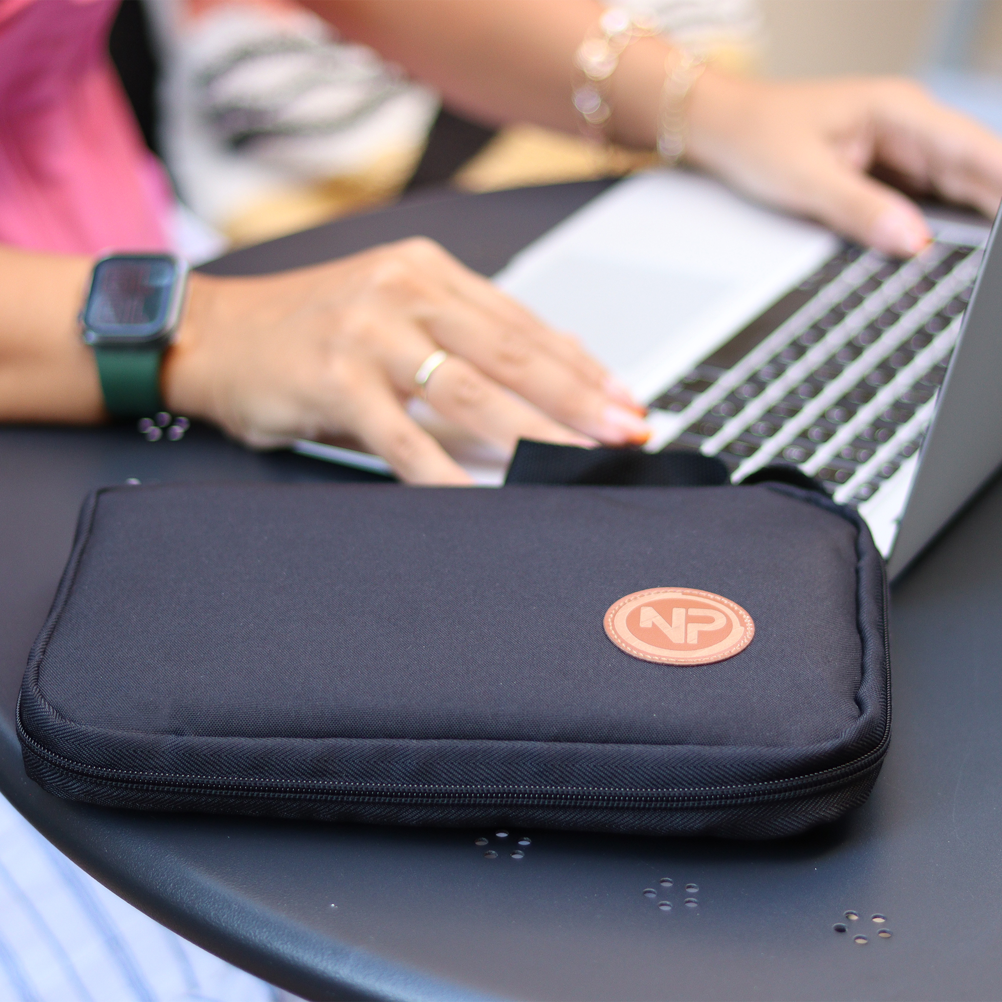 Oval logolu siyah bir çanta, eller laptop kullanırken masanın üzerinde.