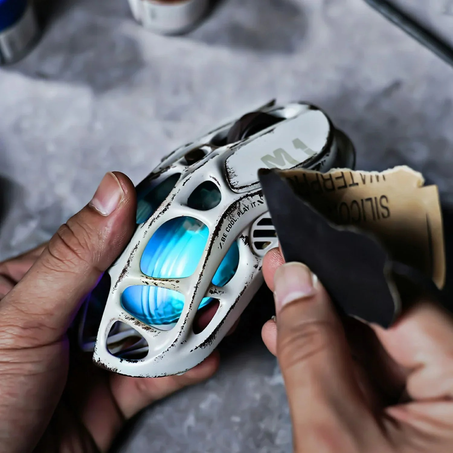 Dłonie trzymają białe, futurystyczne buty z niebieskimi, świecącymi akcentami. Użyto papieru ściernego.