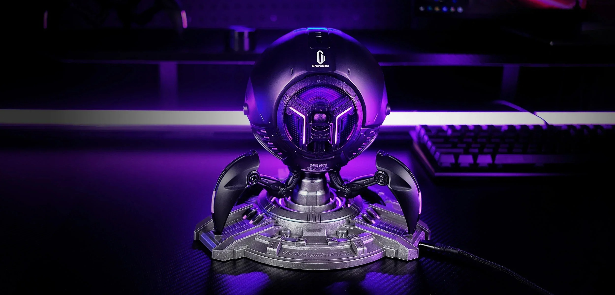 Futurystyczny fioletowy głośnik z mechanicznymi nogami na ciemnym biurku.