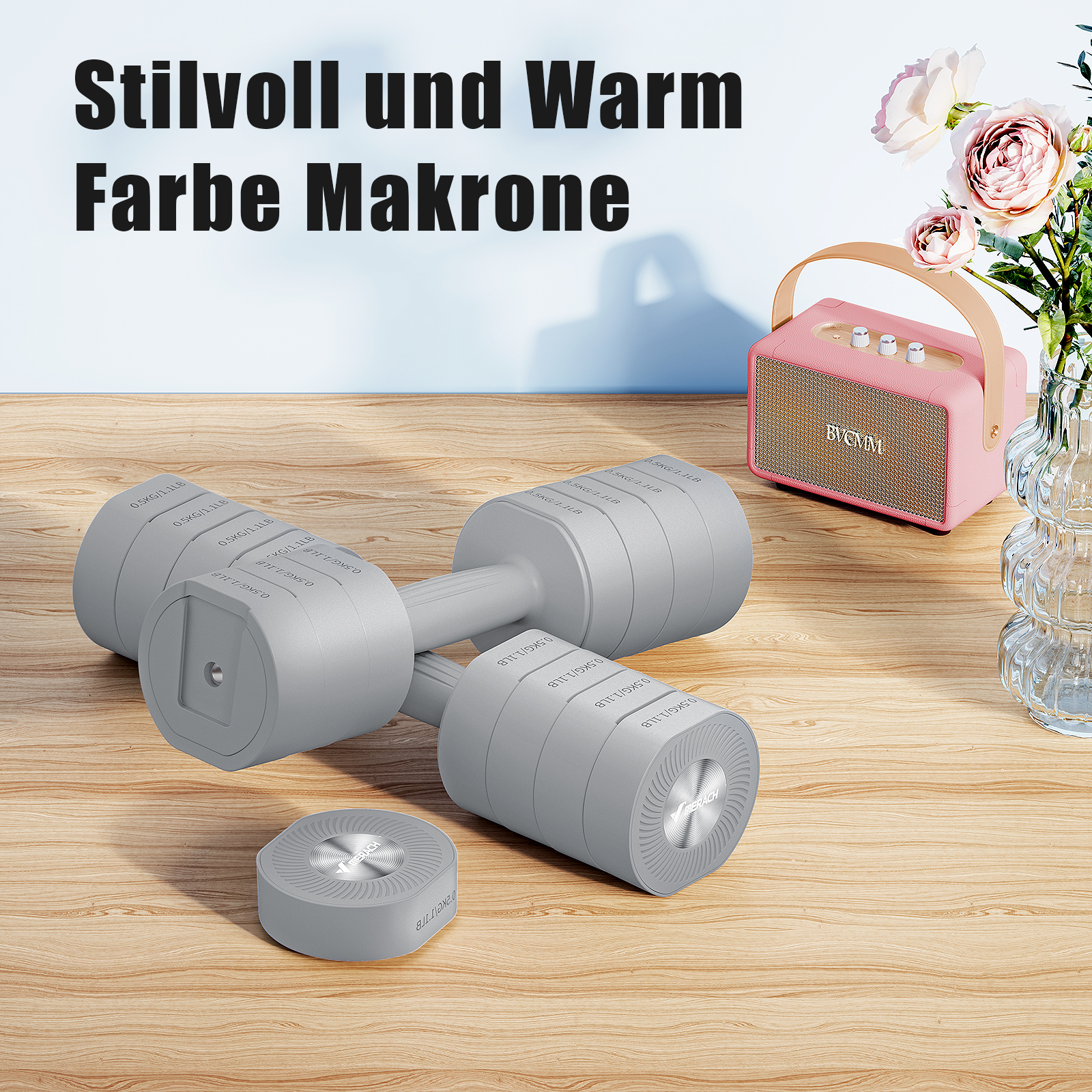Graue Hanteln mit 0,5 kg auf einem Holztisch neben einem rosa Lautsprecher und Blumen.
