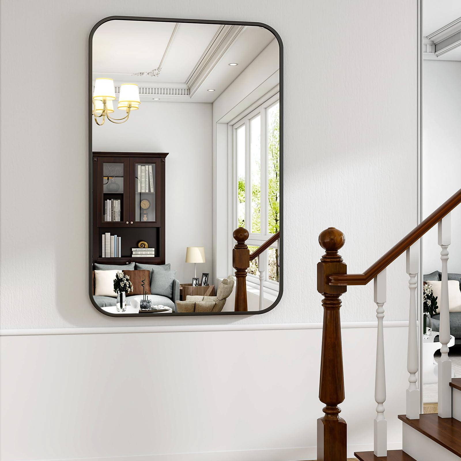 Ein großer schwarzer Spiegel hängt an einer weißen Wand und reflektiert ein Wohnzimmer mit Bücherregal und Sofa.