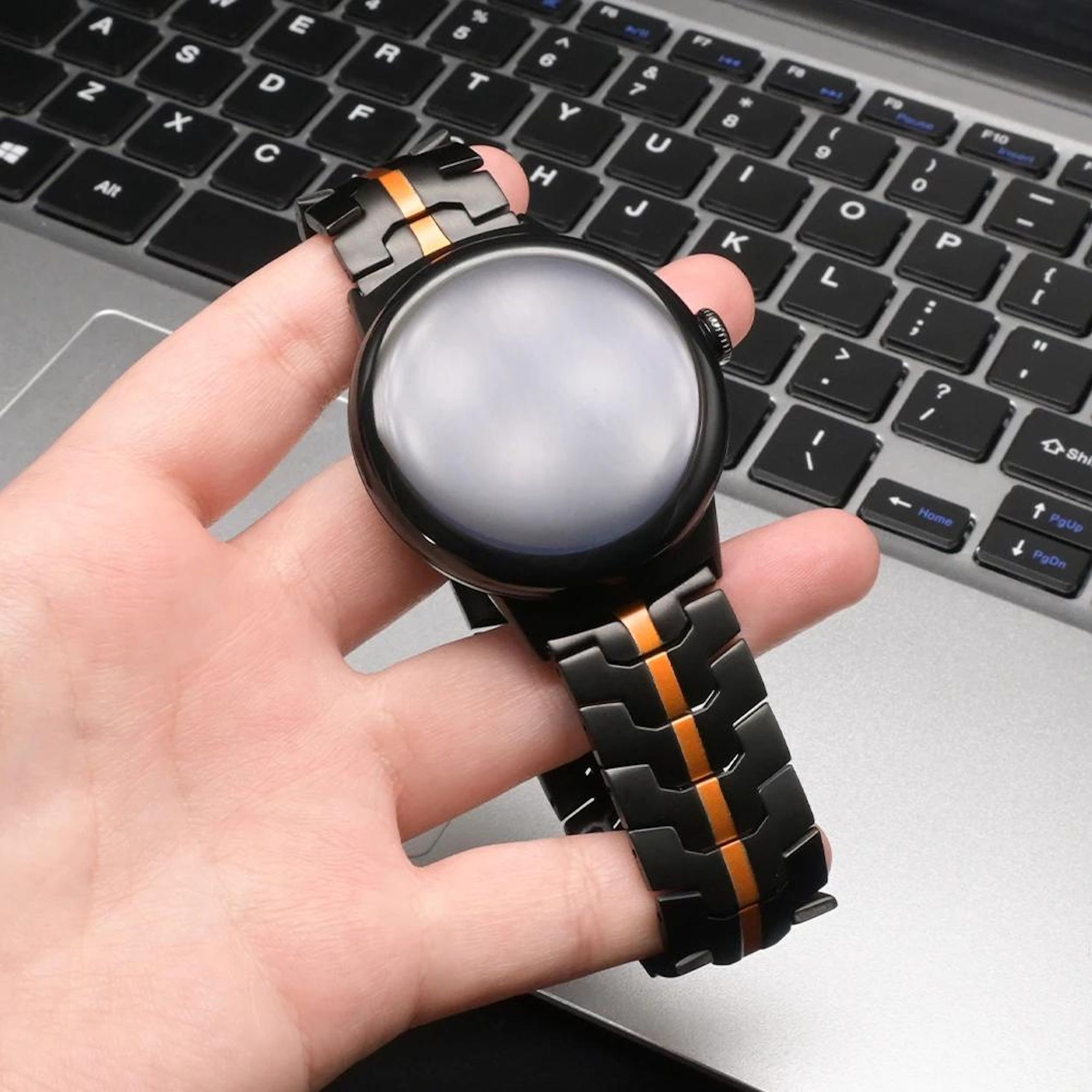Eine Hand hält eine schwarze Uhr mit orangefarbenen Akzenten über einer Laptop-Tastatur.