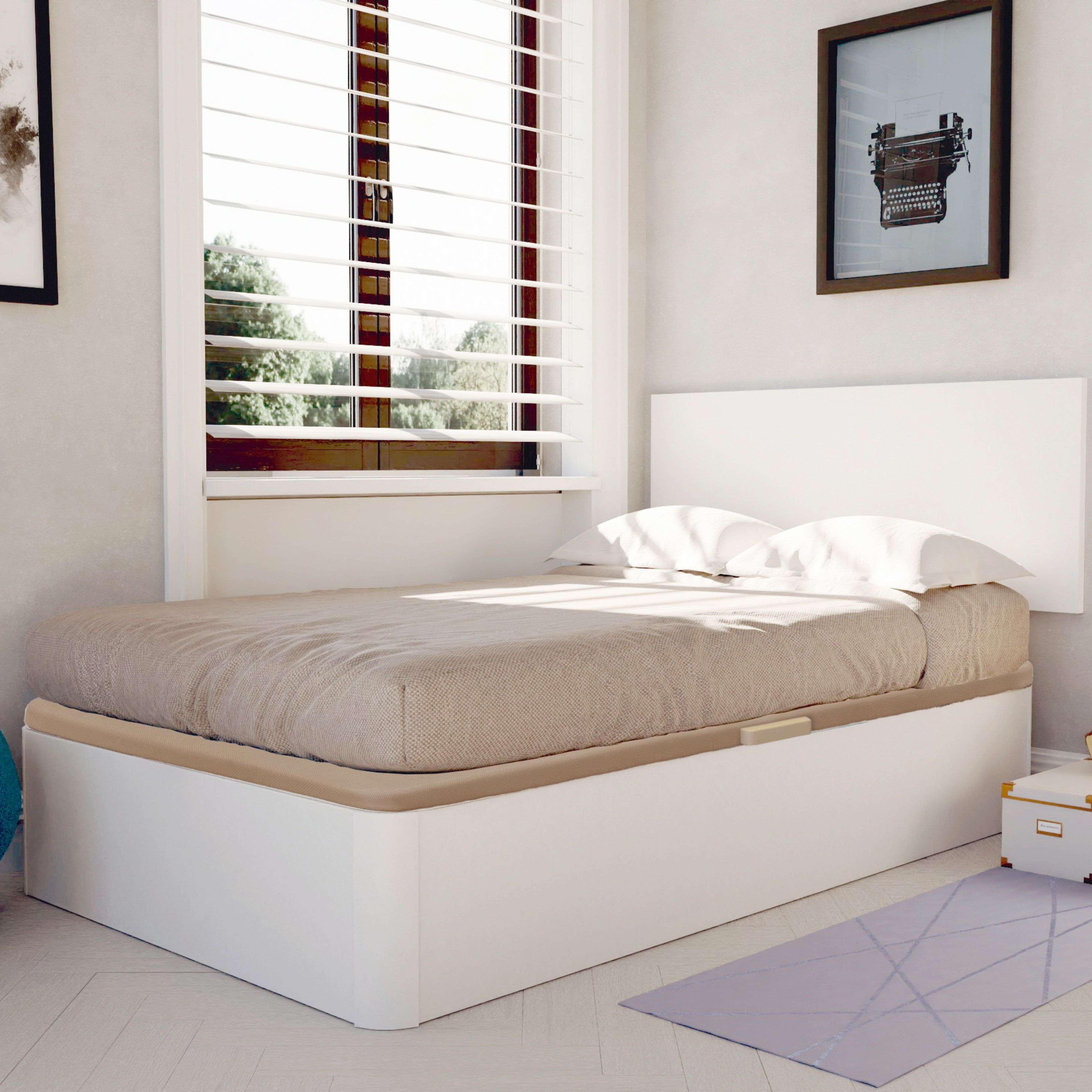 Una cama con manta beige y almohadas blancas está frente a una ventana con persianas.