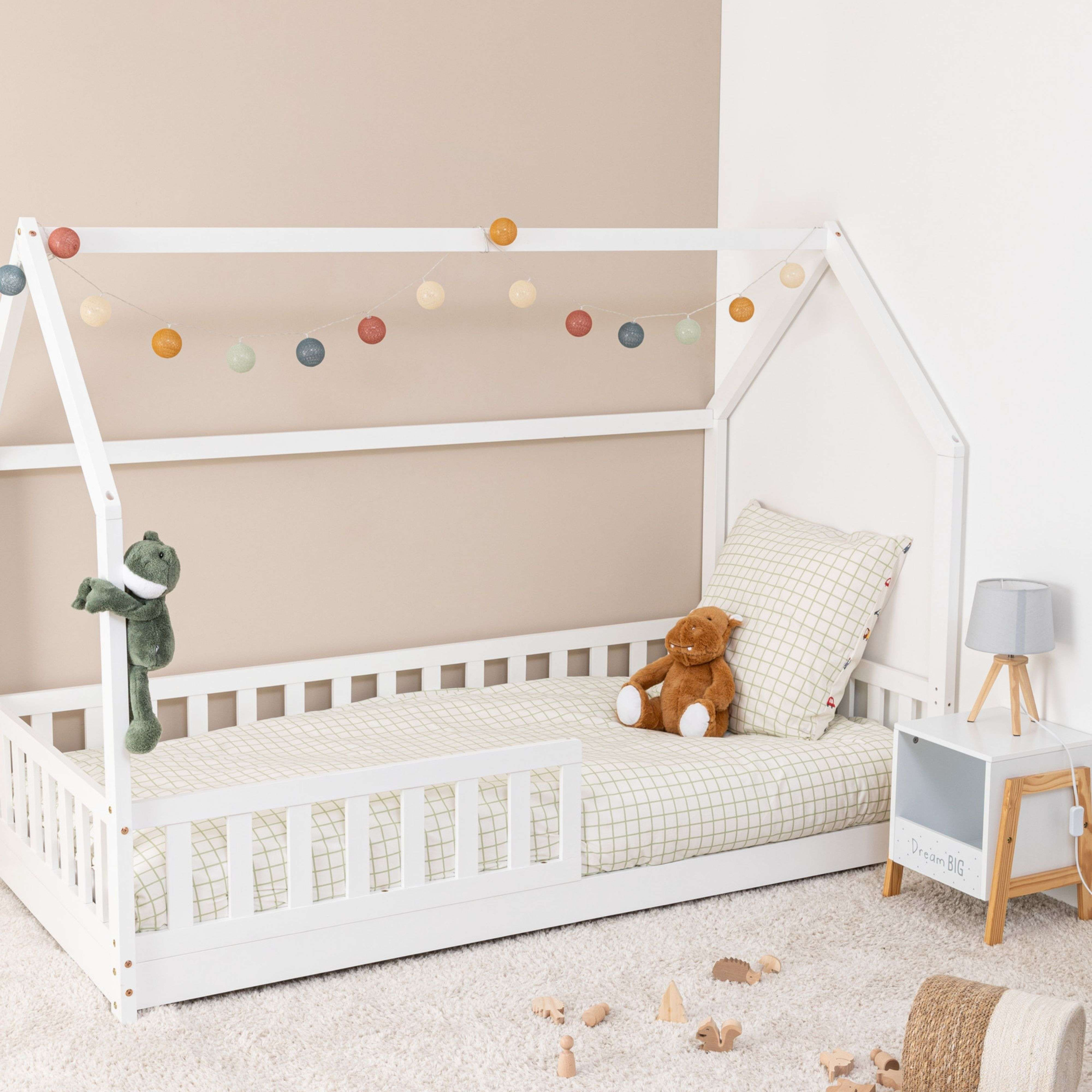 Una cama infantil blanca con luces de colores, juguetes y un peluche de rana.