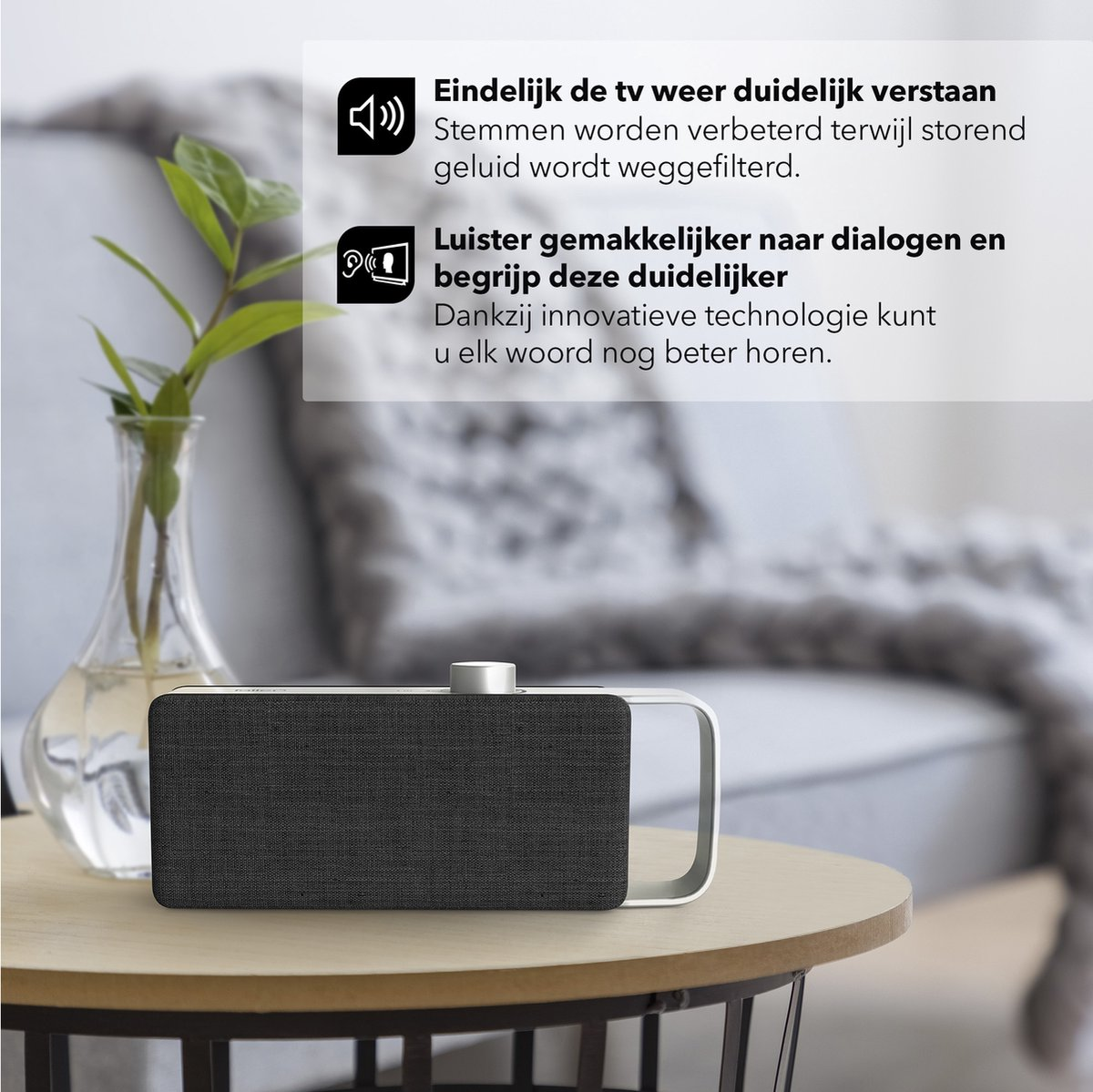 Een zwarte speaker staat op een tafel. Naast de tafel staat een vaas met plant.