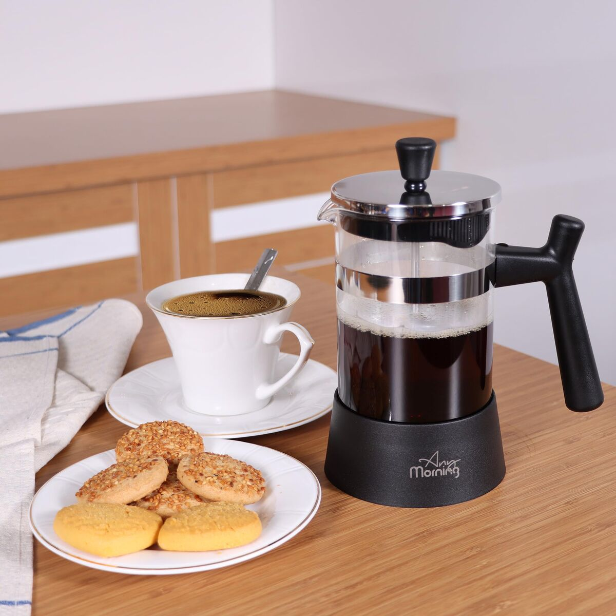 Una taza de café con cuchara, galletas en un plato y una cafetera de prensa con café.