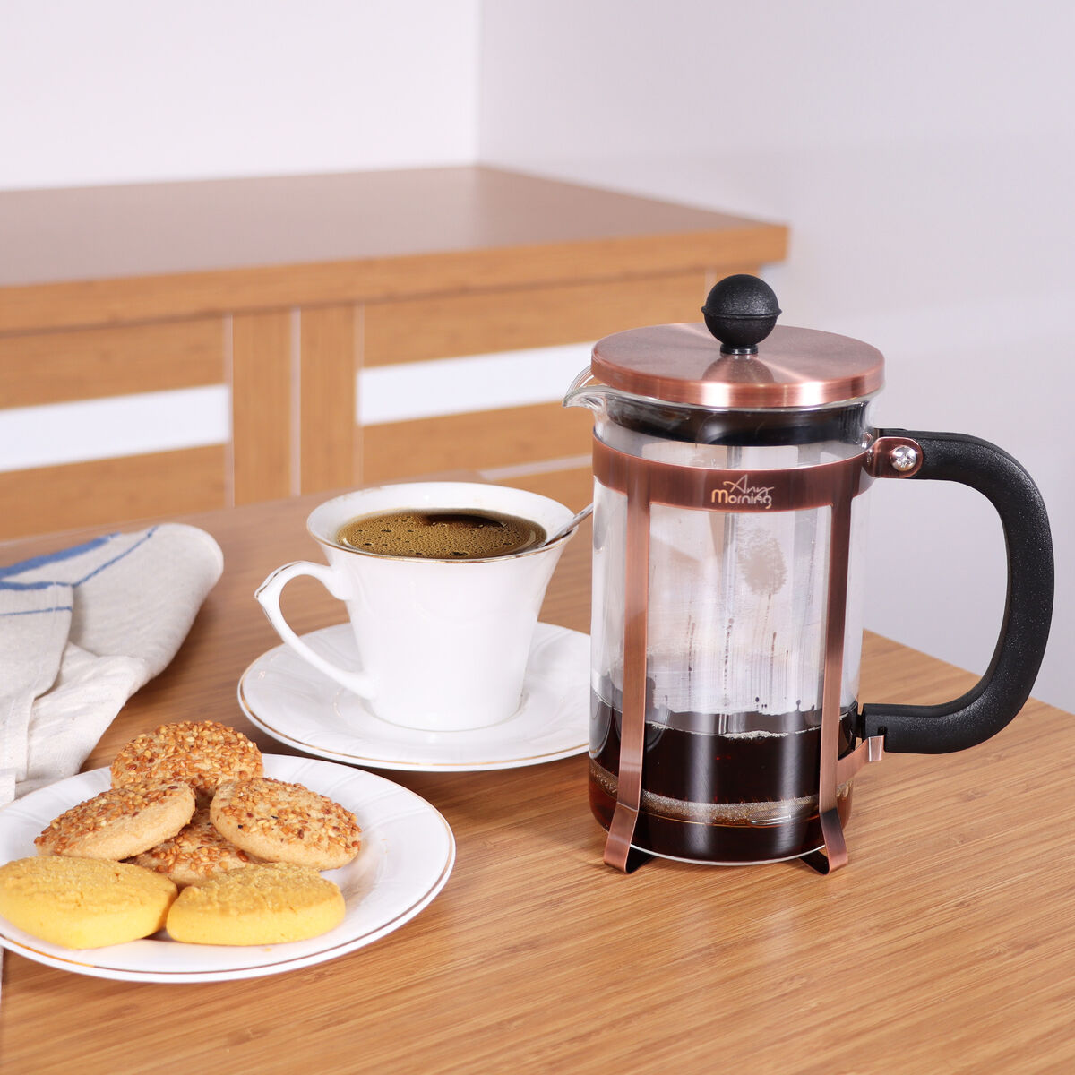 Una prensa francesa de cobre con café, una taza de café blanca y un plato de galletas en una mesa de madera.