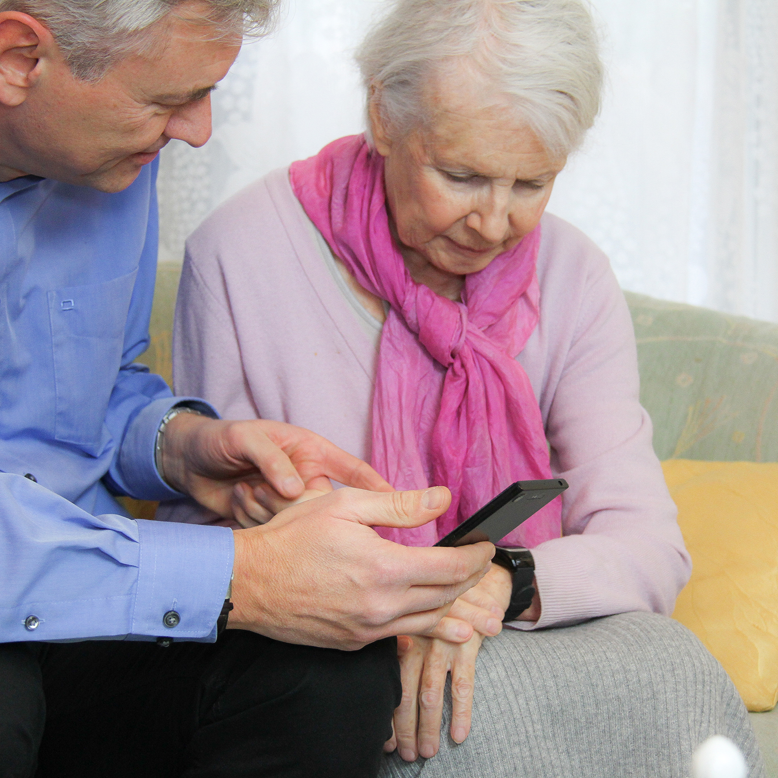 Ein Mann zeigt einer älteren Frau ein Smartphone, während sie auf einem Sofa sitzen.