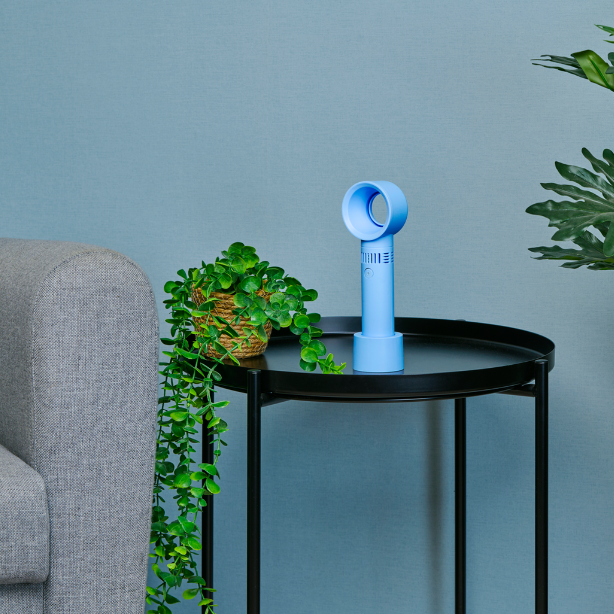 Un ventilateur bleu est posé sur une table noire à côté d'une plante en pot et d'un canapé gris.