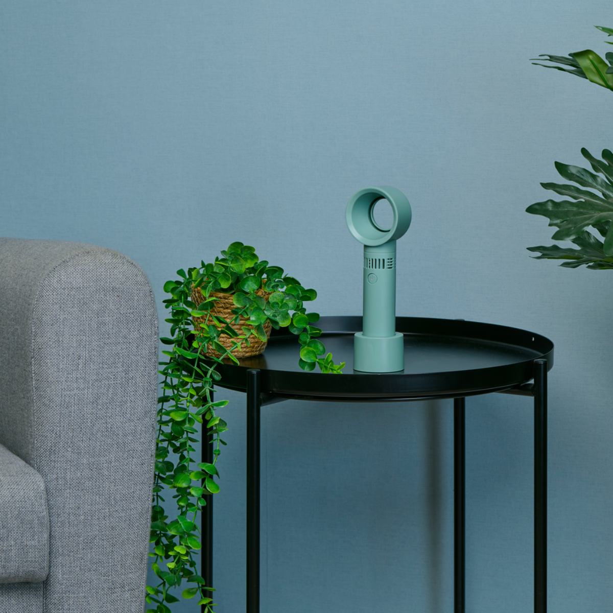 Un ventilateur vert pâle et une plante en pot sur une table noire devant un mur bleu.