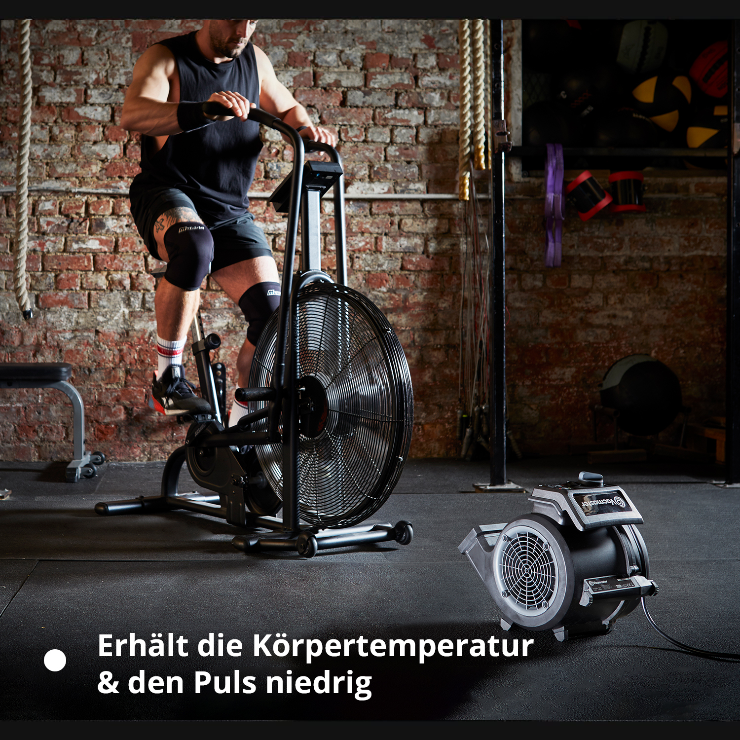 Mann auf Airbike in Turnhalle, mit Ventilator im Vordergrund.