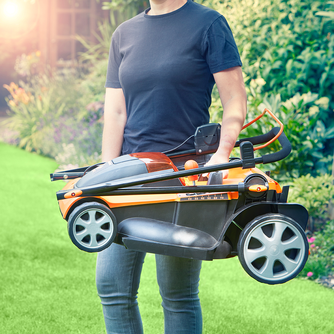 Eine Person trägt einen orangefarbenen Rasenmäher in einem Garten mit grünem Rasen und Blumen.