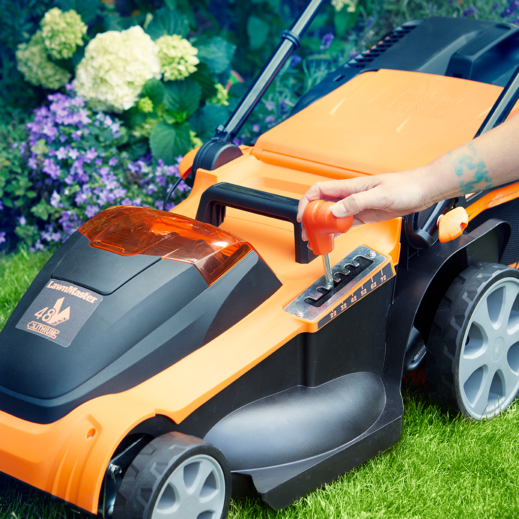 Eine Hand stellt den Hebel an einem orange-schwarzen Rasenmäher ein.