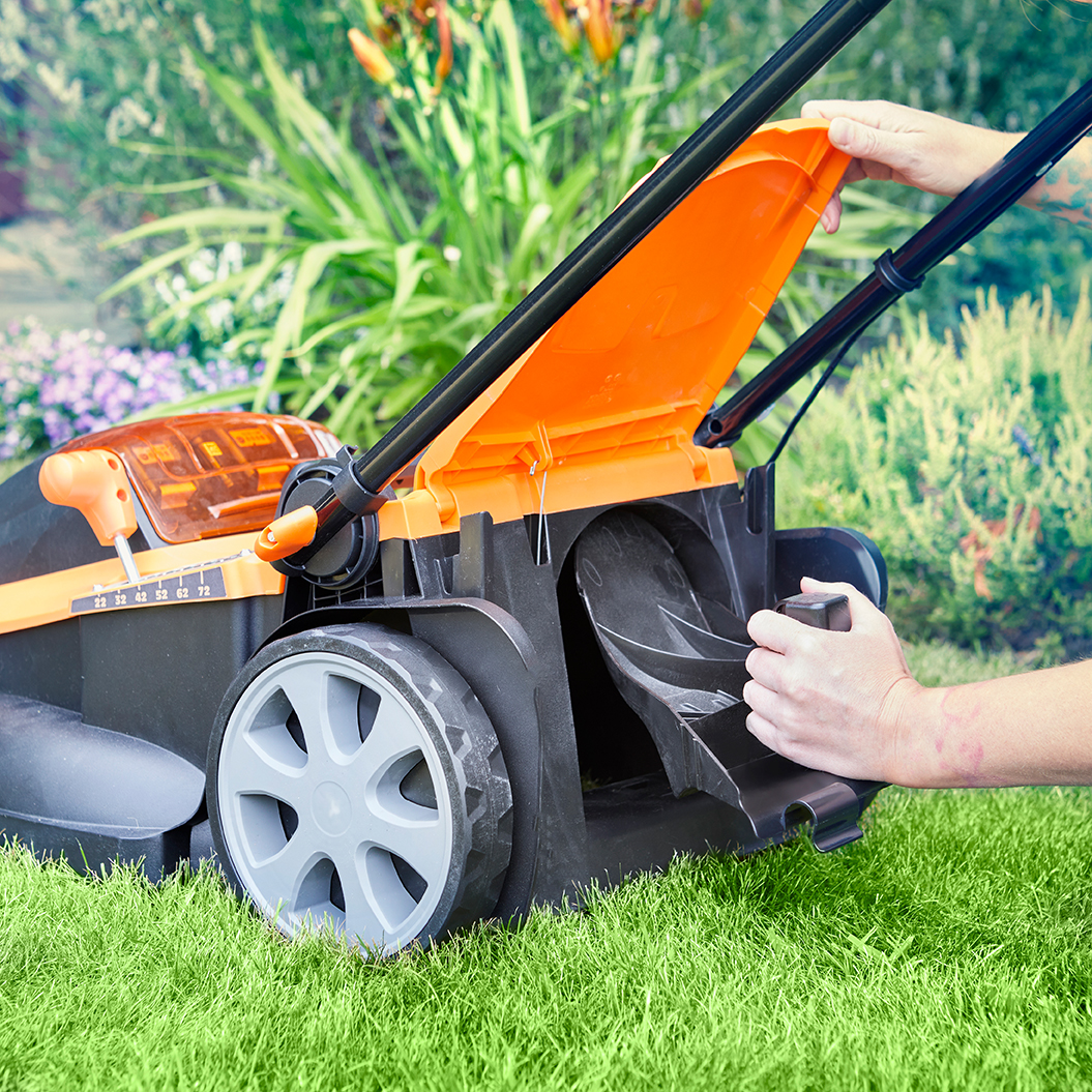 Person öffnet orangefarbenen Deckel eines Rasenmähers im Garten auf grünem Gras.