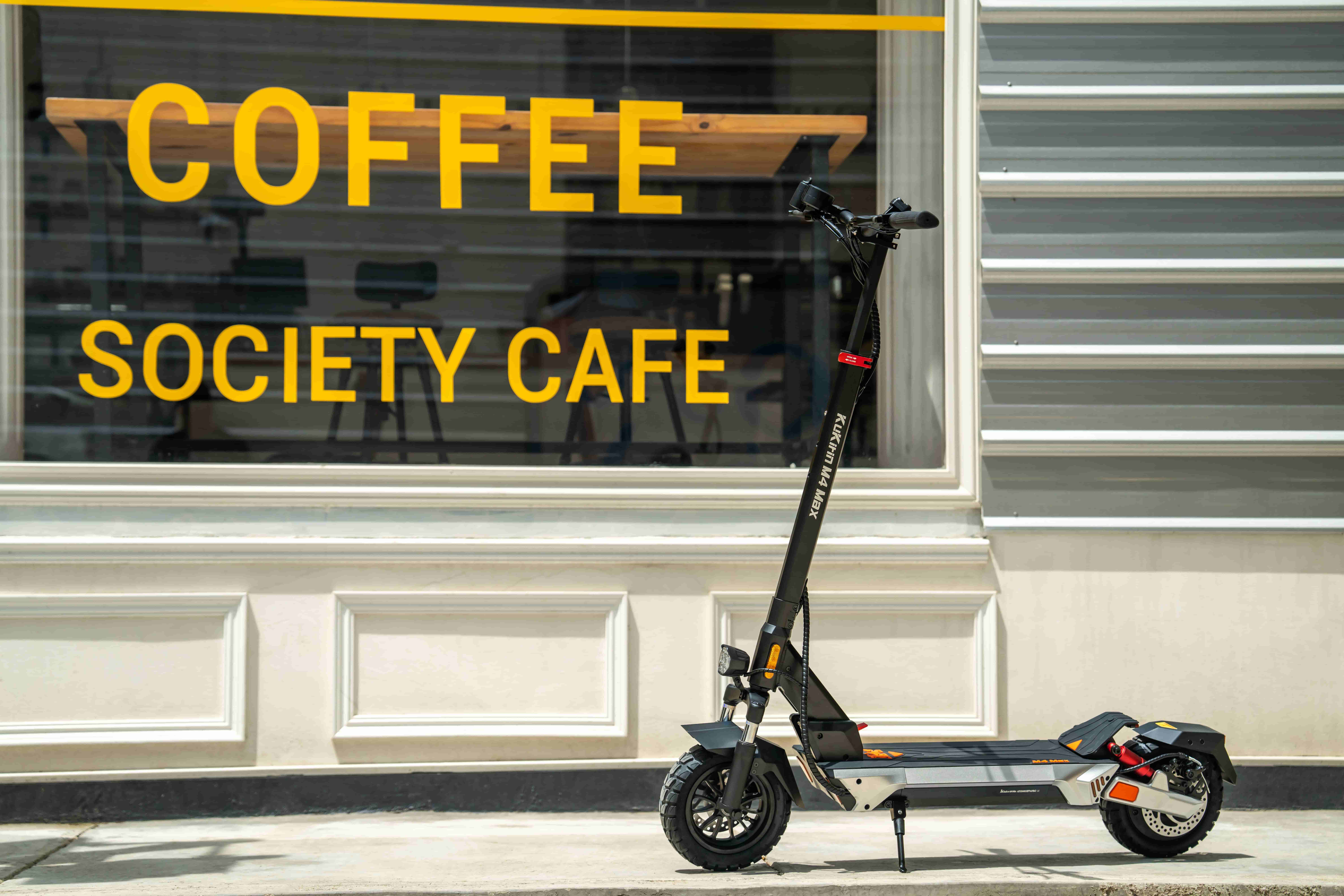 Ein schwarzer Elektroroller steht vor einem Café mit gelben Schriftzügen "COFFEE SOCIETY CAFE".