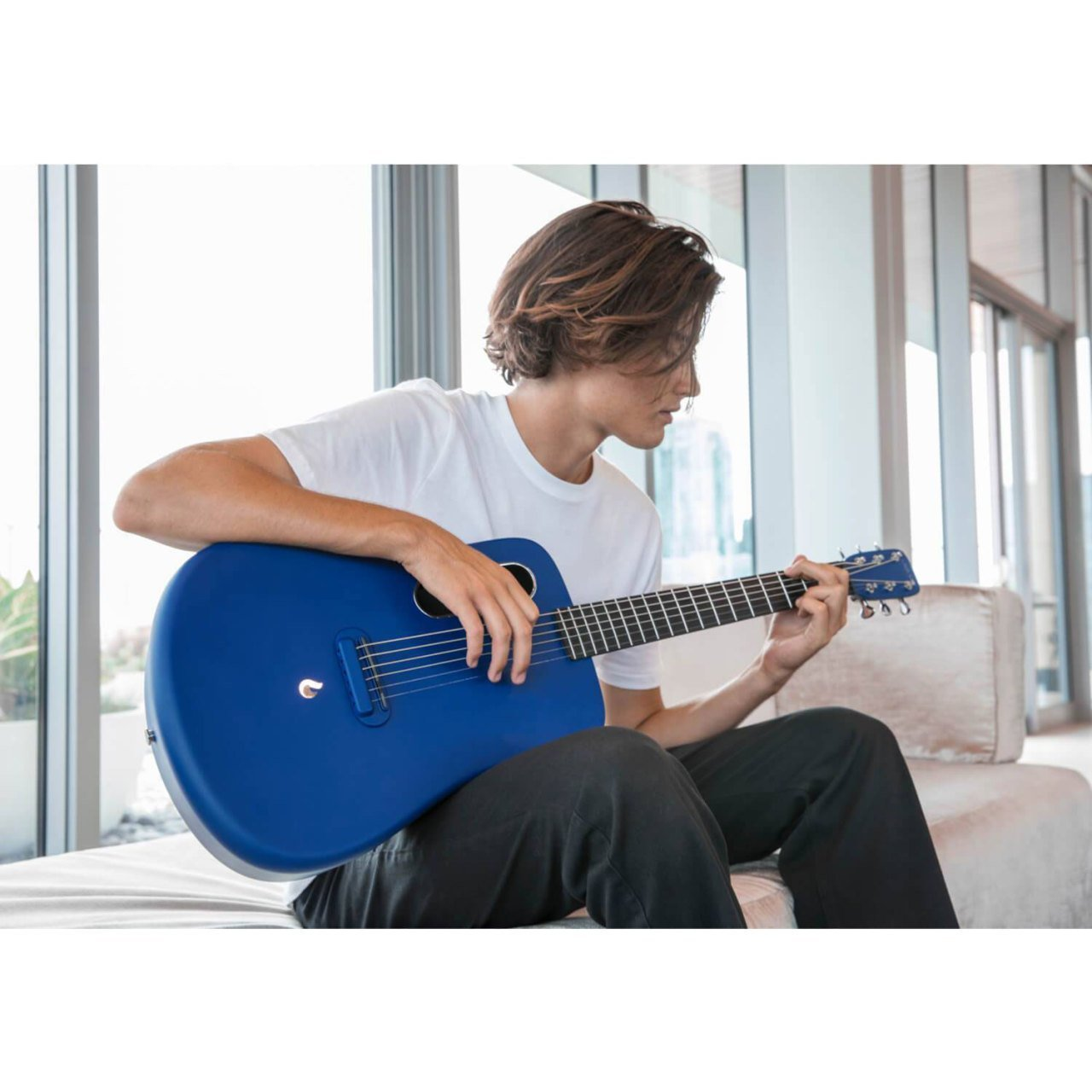 Beyaz tişörtlü genç bir adam koltukta otururken parlak mavi gitar çalıyor.