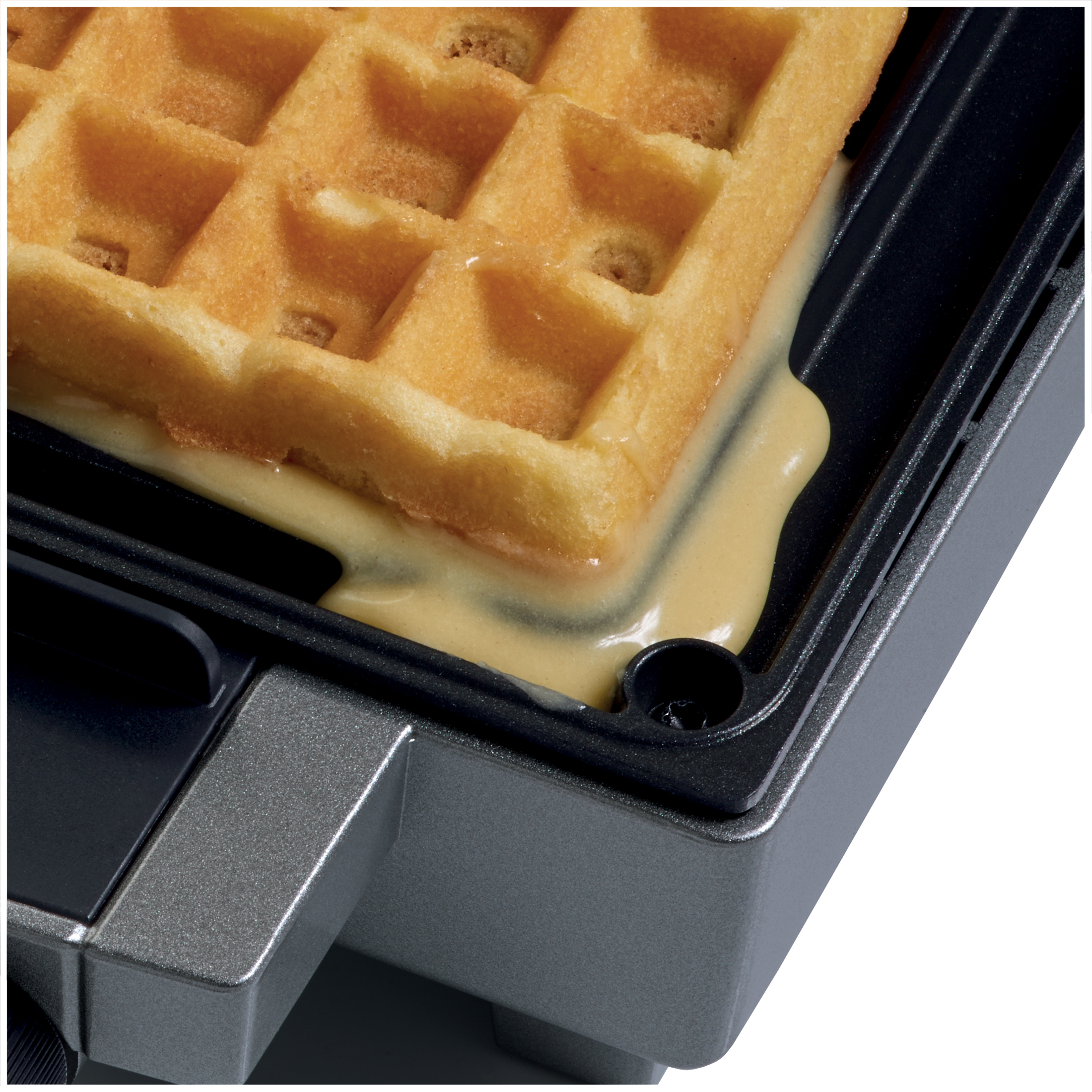 A golden waffle is cooking in a dark waffle maker, with batter spilling over the edge.